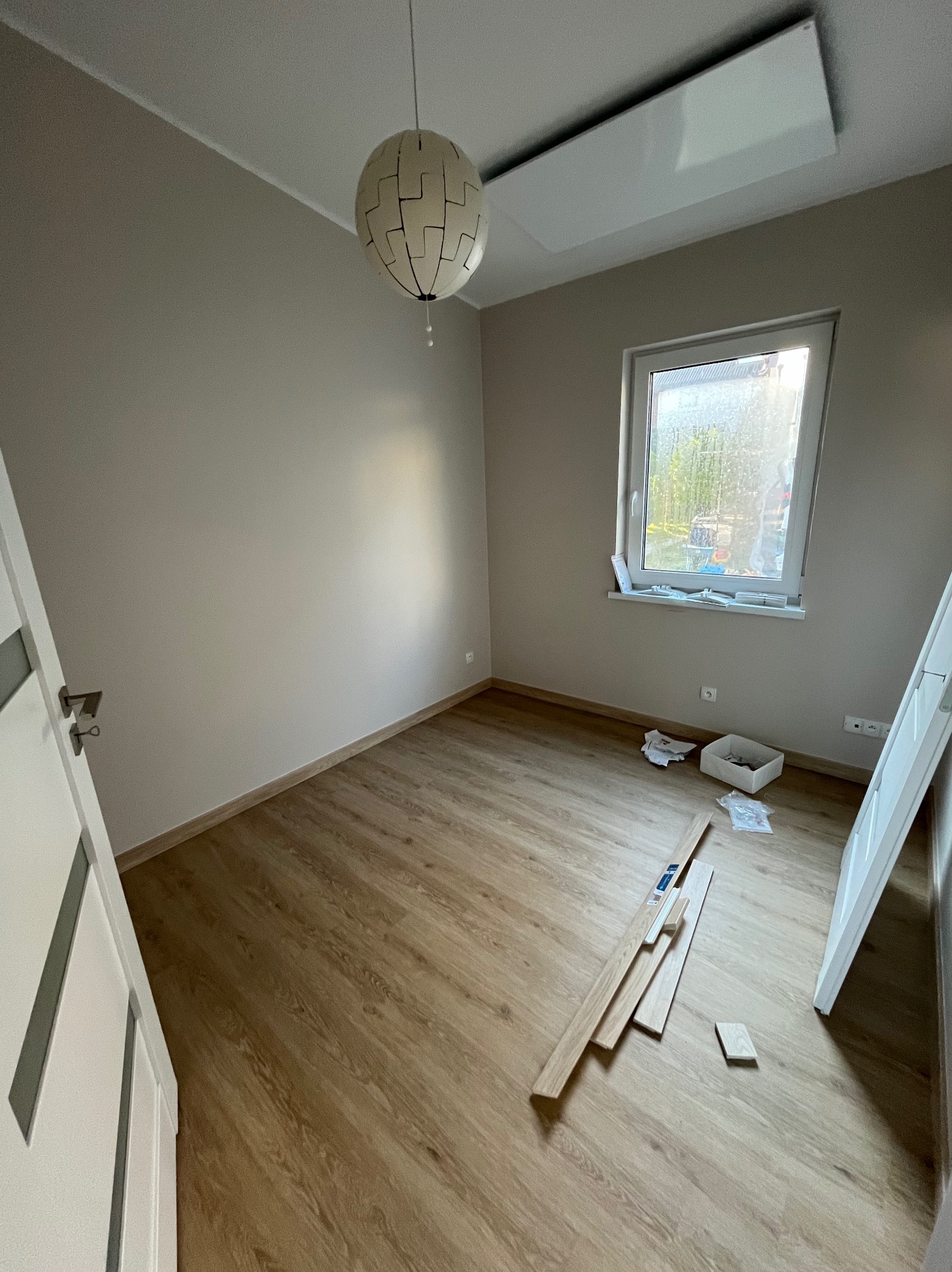 Wnętrze pokoju w trakcie prac wykończeniowych: panele podłogowe częściowo ułożone, widoczne deski paneli na podłodze, biała lampa wisząca o ażurowym kloszu, jasne ściany i białe drzwi z szarymi...