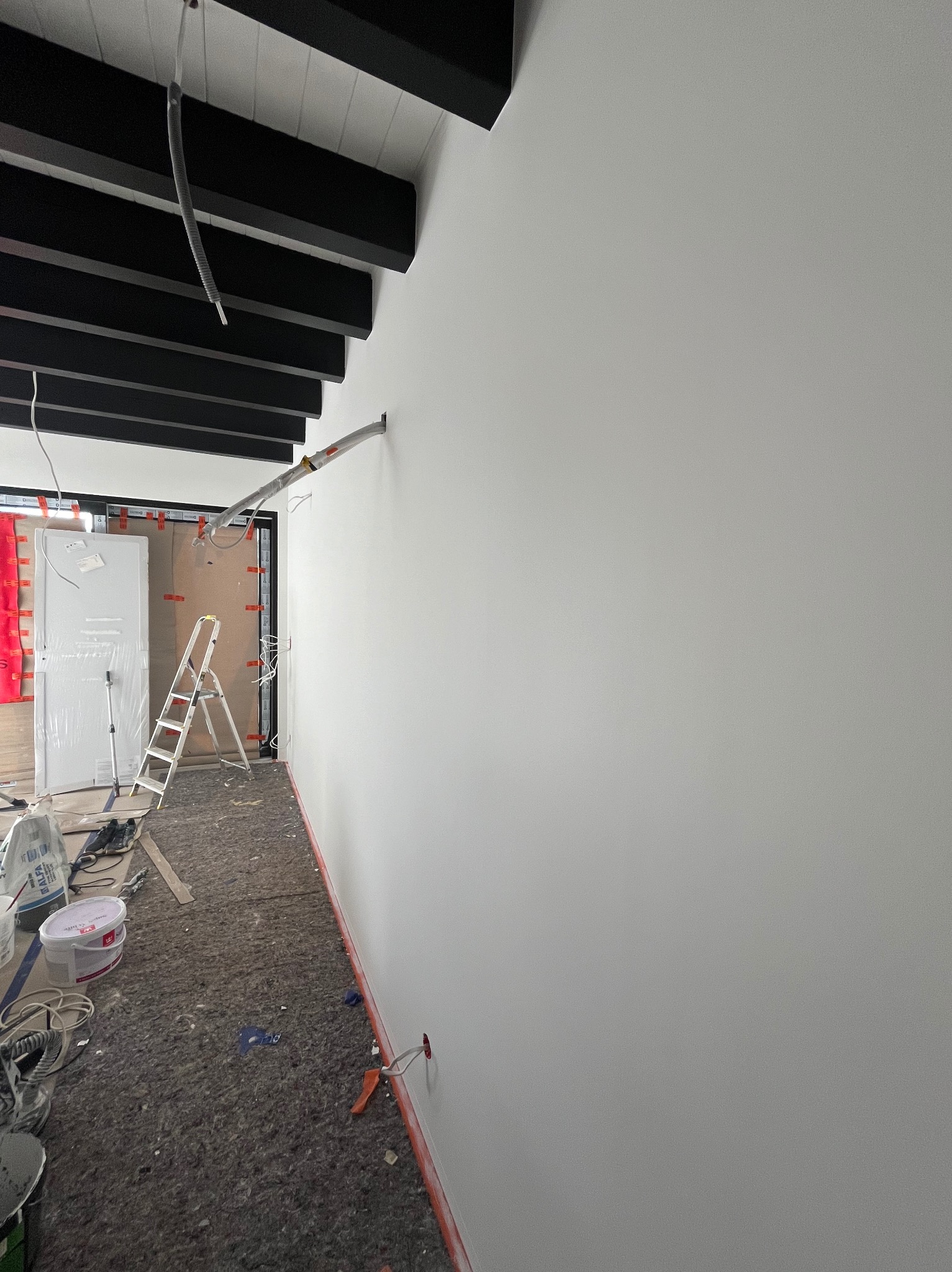 Wnętrze w trakcie remontu, ściana pomalowana na biało, widoczne przewody, drabina, wiaderka z farbą i zabezpieczona podłoga.