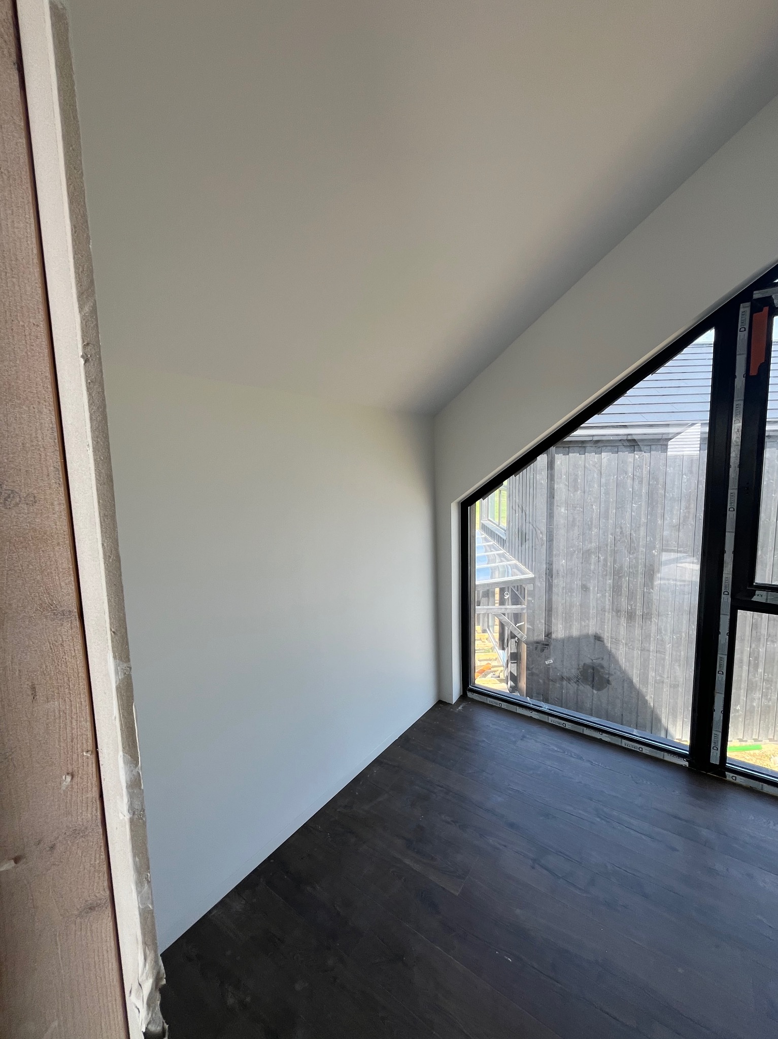 Wnętrze pokoju z ukośną ścianą i oknem o nieregularnym kształcie, widoczne świeżo położone panele podłogowe w ciemnym kolorze, ściany pomalowane na biało, na jednej ze ścian widoczny surowy...