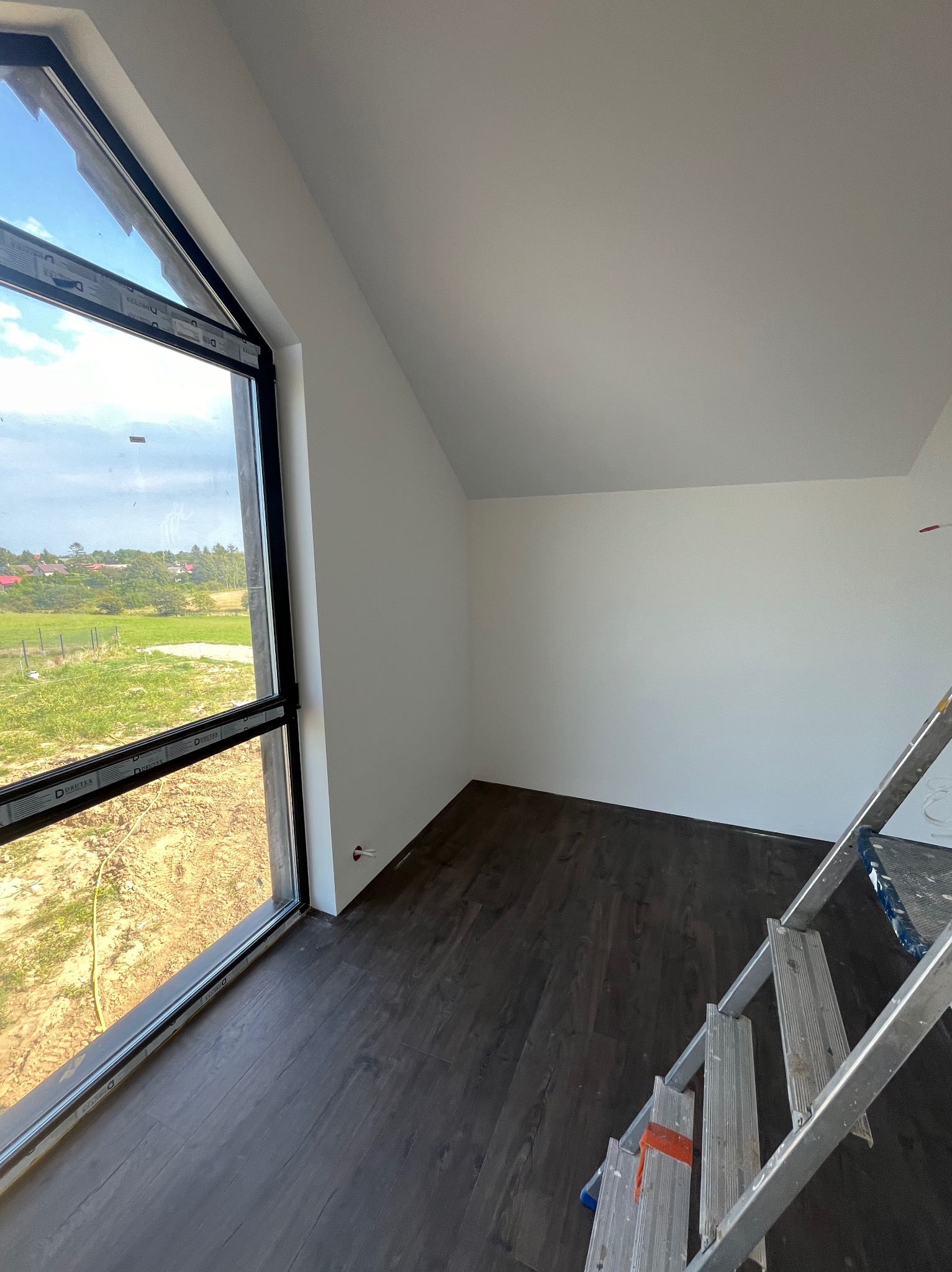Pokój na poddaszu z oknem trapezowym i ciemnymi panelami podłogowymi, ściany pomalowane na biało, widoczna drabina malarska.