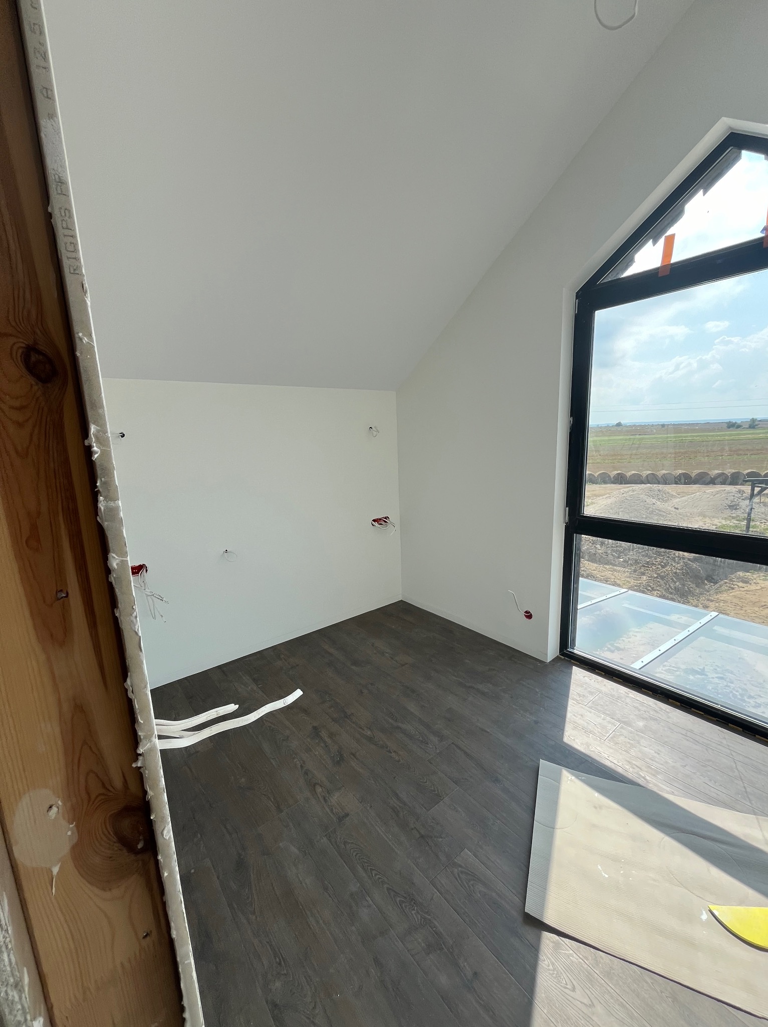 Pokój na poddaszu z panelami podłogowymi w trakcie układania, widoczne fragmenty paneli, białe ściany z wyprowadzonymi przewodami elektrycznymi, okno z widokiem na pole, fragment ościeżnicy...