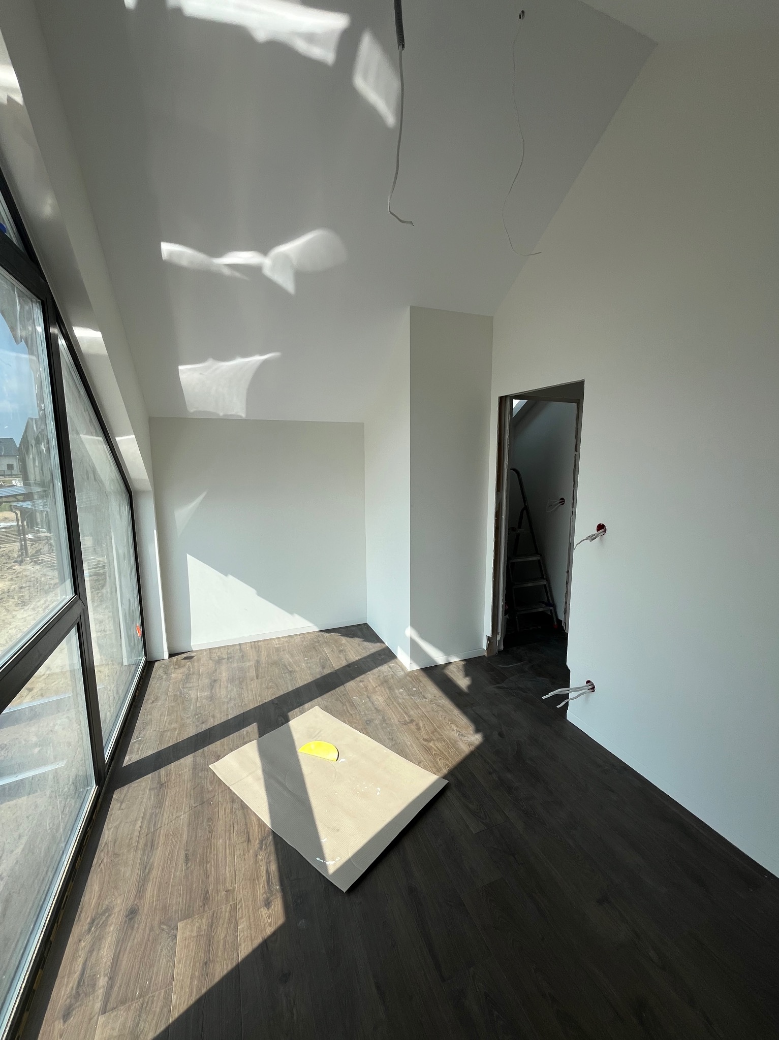 Jasne wnętrze w stanie deweloperskim z dużym oknem, panelami na podłodze i świeżo pomalowanymi ścianami na biało. Na podłodze leży karton ochronny z żółtą plamą. W głębi widoczne otwarte drzwi...