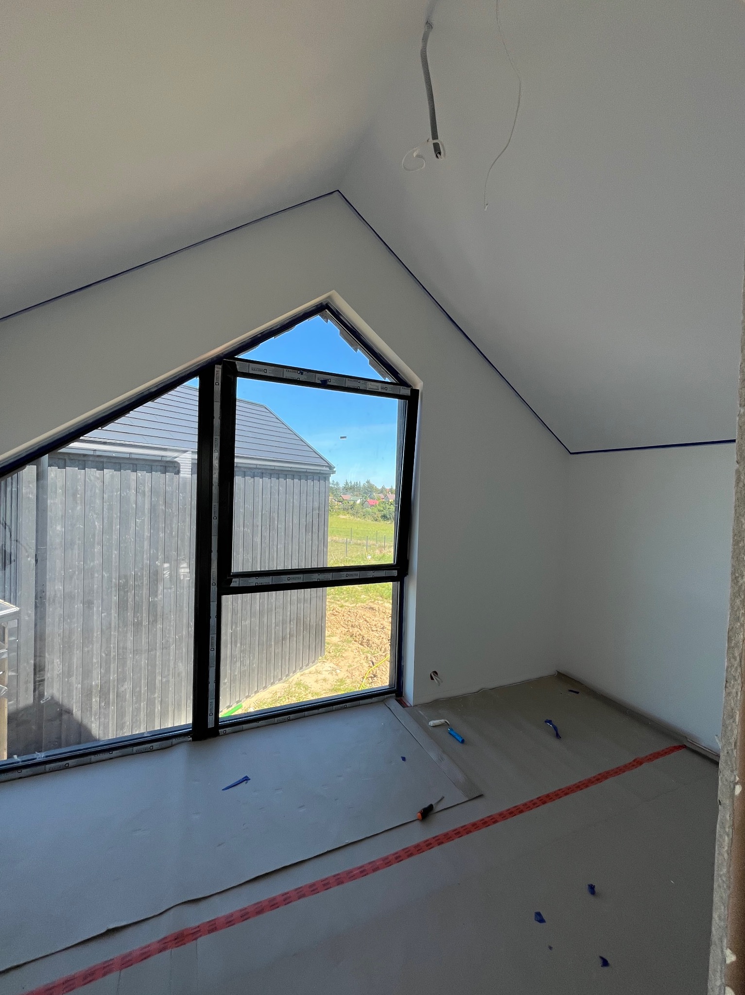 Wnętrze poddasza z oknem w kształcie trapezu, ściany przygotowane do malowania, zabezpieczone taśmą malarską, podłoga zabezpieczona folią, widoczne narzędzia malarskie.