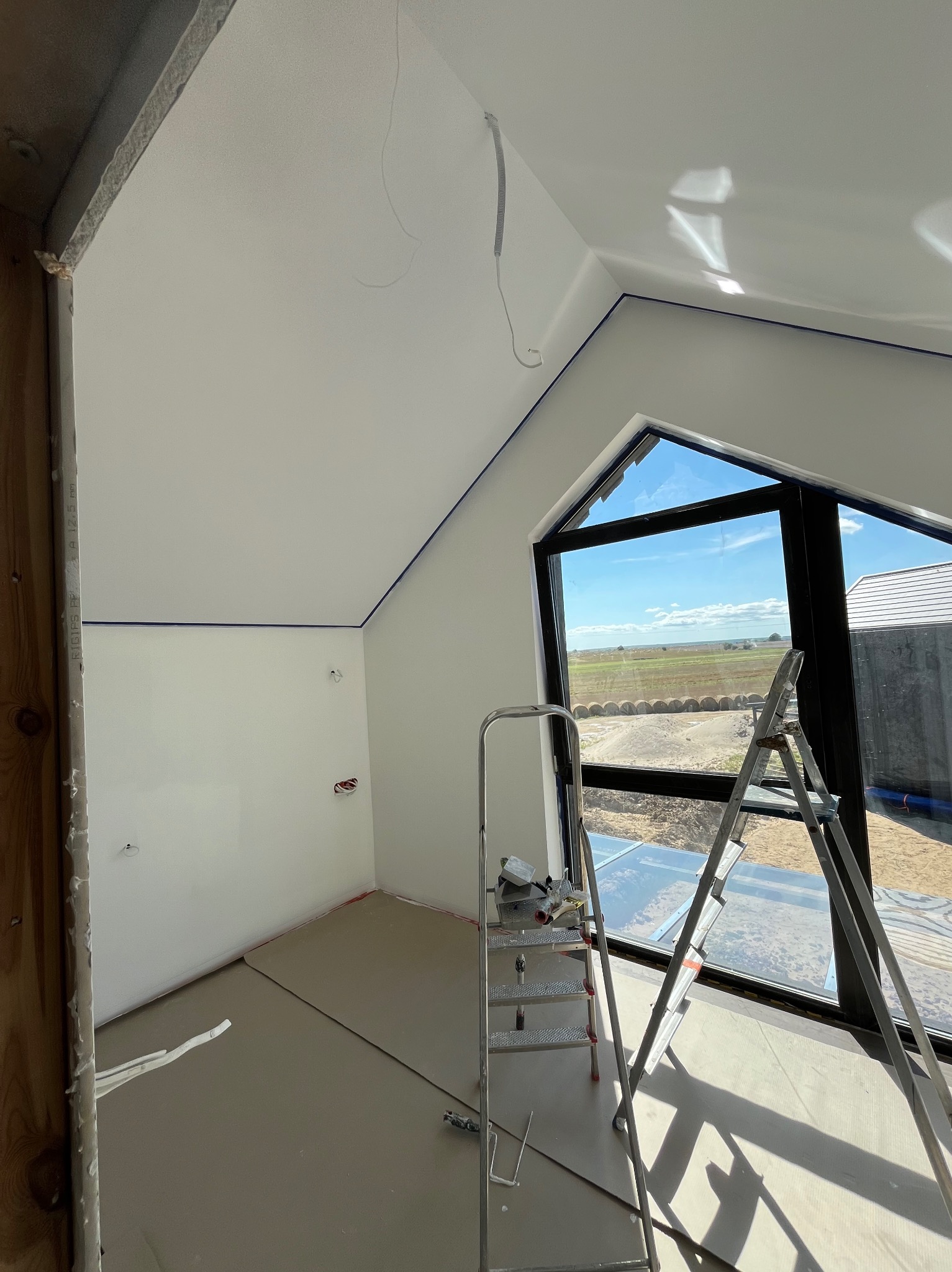 Pokój na poddaszu w trakcie prac wykończeniowych, ściany i sufit pomalowane na biało, zabezpieczone niebieską taśmą malarską, widoczna drabina aluminiowa i narzędzia malarskie, okno z widokiem...