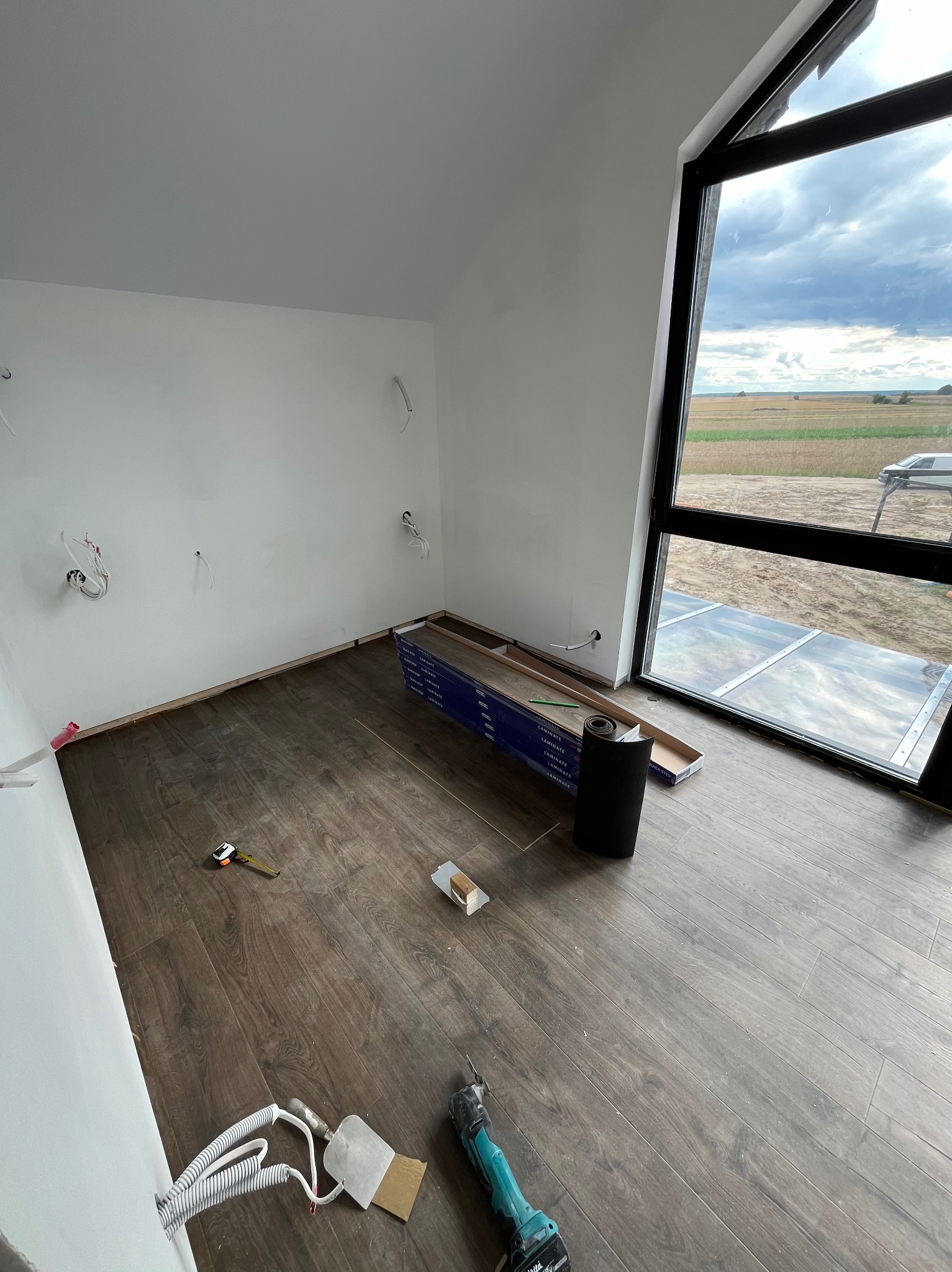 Pokój w trakcie remontu z częściowo ułożoną podłogą z paneli, widoczne narzędzia, rolka podkładu i paczki paneli przy oknie z widokiem na pole.