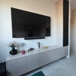 Nowoczesna szafka RTV z szarymi frontami, dekoracjami i dużym telewizorem na ścianie. Obok zabudowa z czarnym panelem. Marmurowe płytki na podłodze.