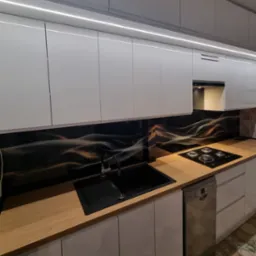 Nowoczesna kuchnia z białymi szafkami, drewnianym blatem i czarnym panelem ze złotym wzorem. Widoczny zlew, płyta grzewcza i oświetlenie LED pod szafkami.