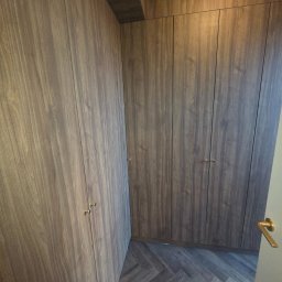 Om MEBLE NA WYMIAR - Wnętrze z zabudową szafy wnękowej z drewnopodobnymi frontami i drobnymi, złotymi uchwytami. Na podłodze parkiet ułożony w jodełkę.