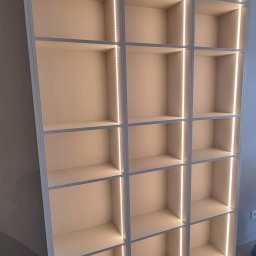 Om MEBLE NA WYMIAR - Biała, nowoczesna szafa wnękowa z podświetleniem LED, widok z dołu. Półki puste, minimalistyczny design, ciepłe światło podkreśla geometrię.