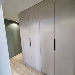 Om MEBLE NA WYMIAR - Jasnoszara szafa wnękowa z minimalistycznymi, czarnymi uchwytami, wkomponowana w korytarz z drewnianą podłogą i zieloną ścianą w tle.