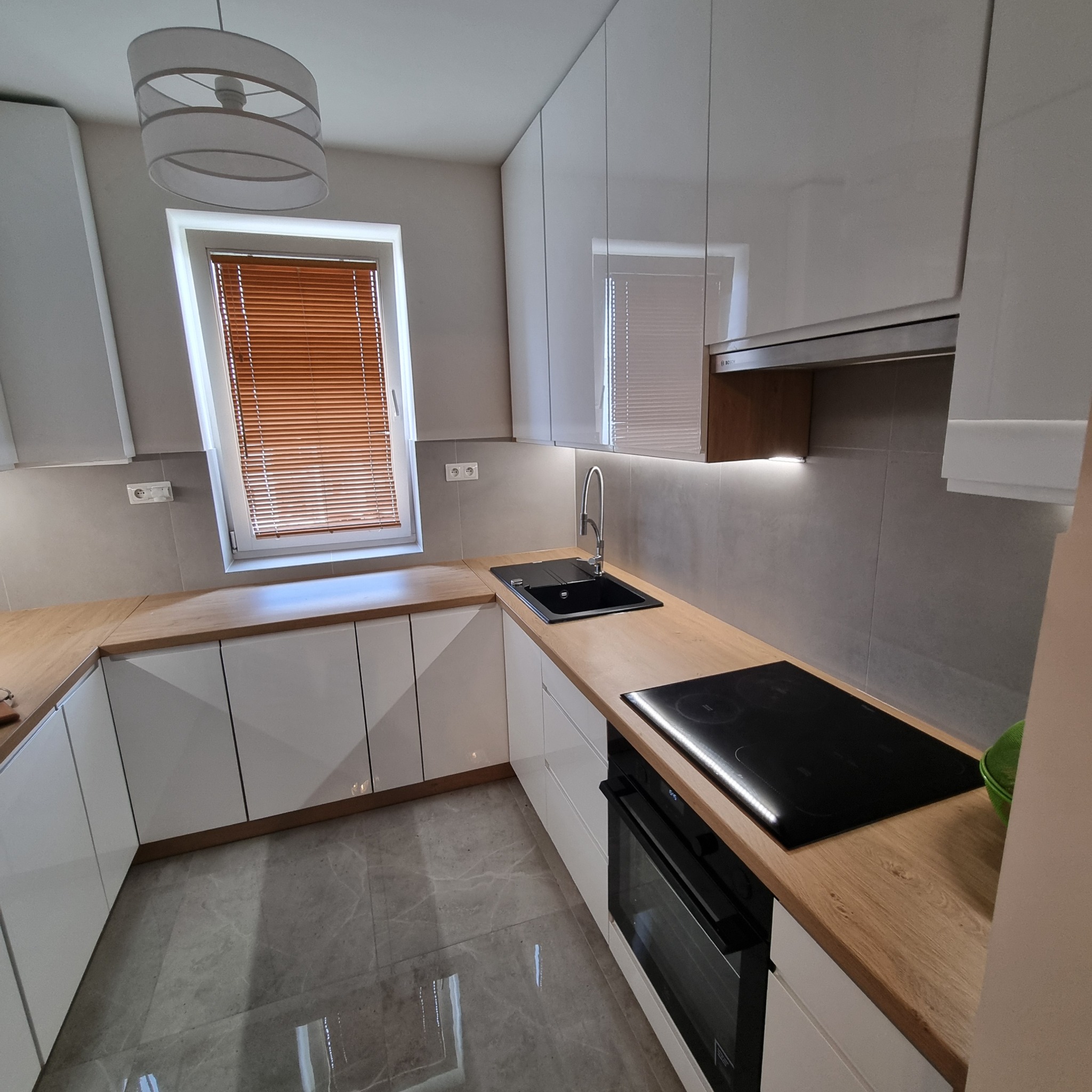 Nowoczesna kuchnia z białymi, lakierowanymi frontami, drewnianym blatem i szarą glazurą. Widoczny zlew, płyta indukcyjna i okno z brązowymi żaluzjami.