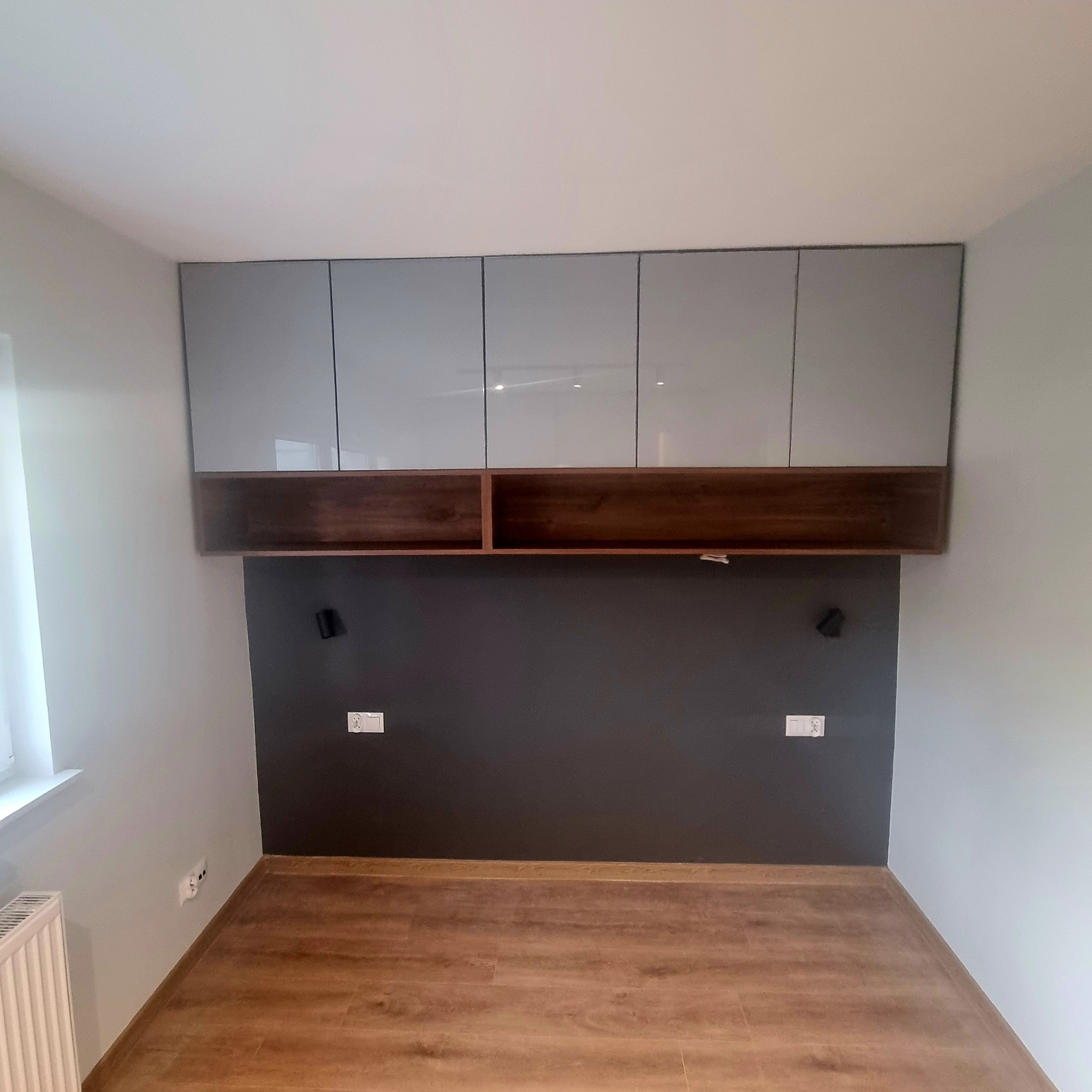 Szafa wnękowa z szarymi, lakierowanymi frontami i drewnianą półką nad ciemną ścianą w pokoju. Widoczne gniazdka elektryczne i podłoga z paneli.