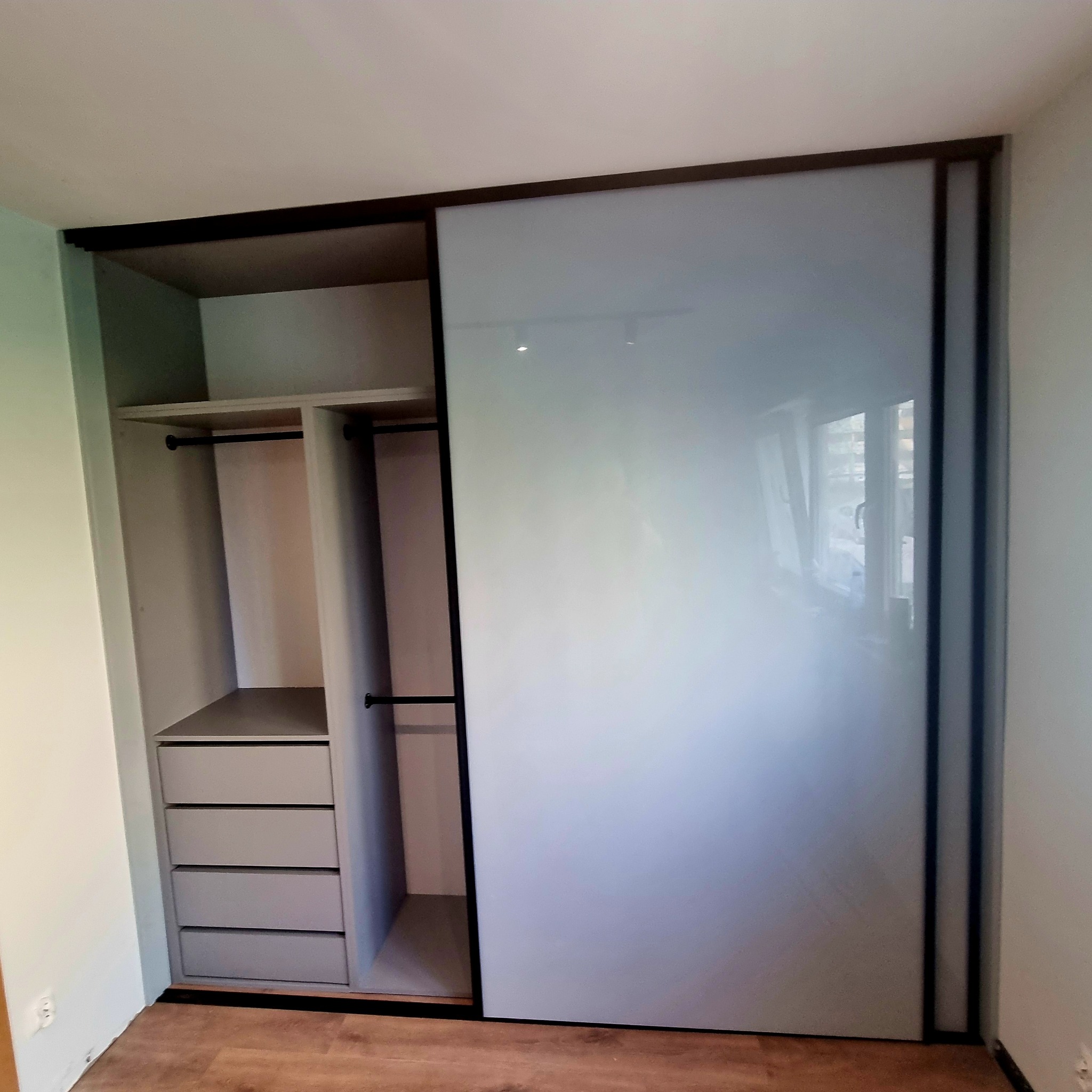 Szafa wnękowa z przesuwnymi drzwiami w odcieniach szarości. Widoczny system półek i szuflad wewnątrz. Minimalistyczny design z ciemnymi obramowaniami.