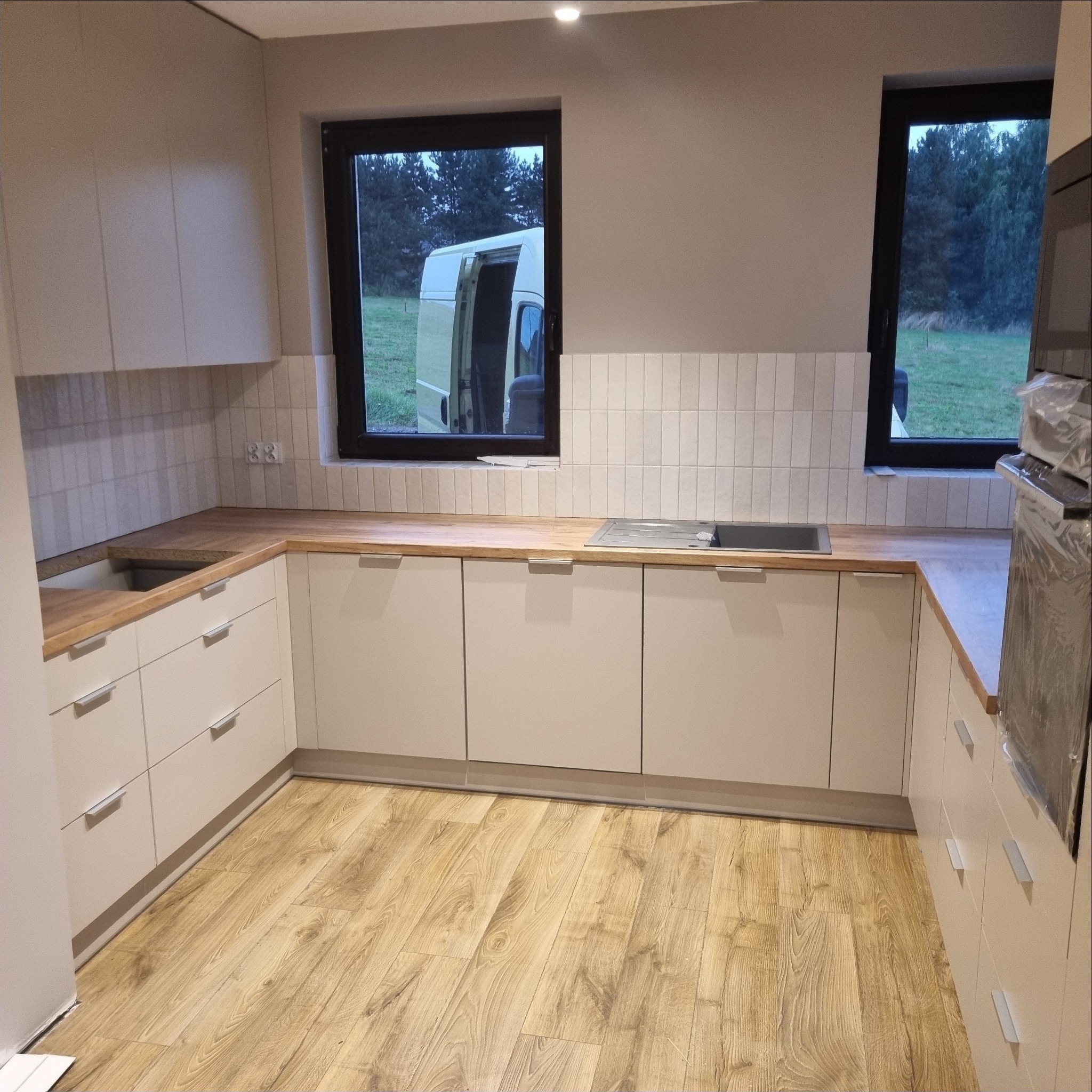 Jasna kuchnia z drewnianym blatem i białymi frontami szafek, widok na zewnątrz przez okno z dostawczym samochodem. Minimalistyczny styl, płytki nad blatem.