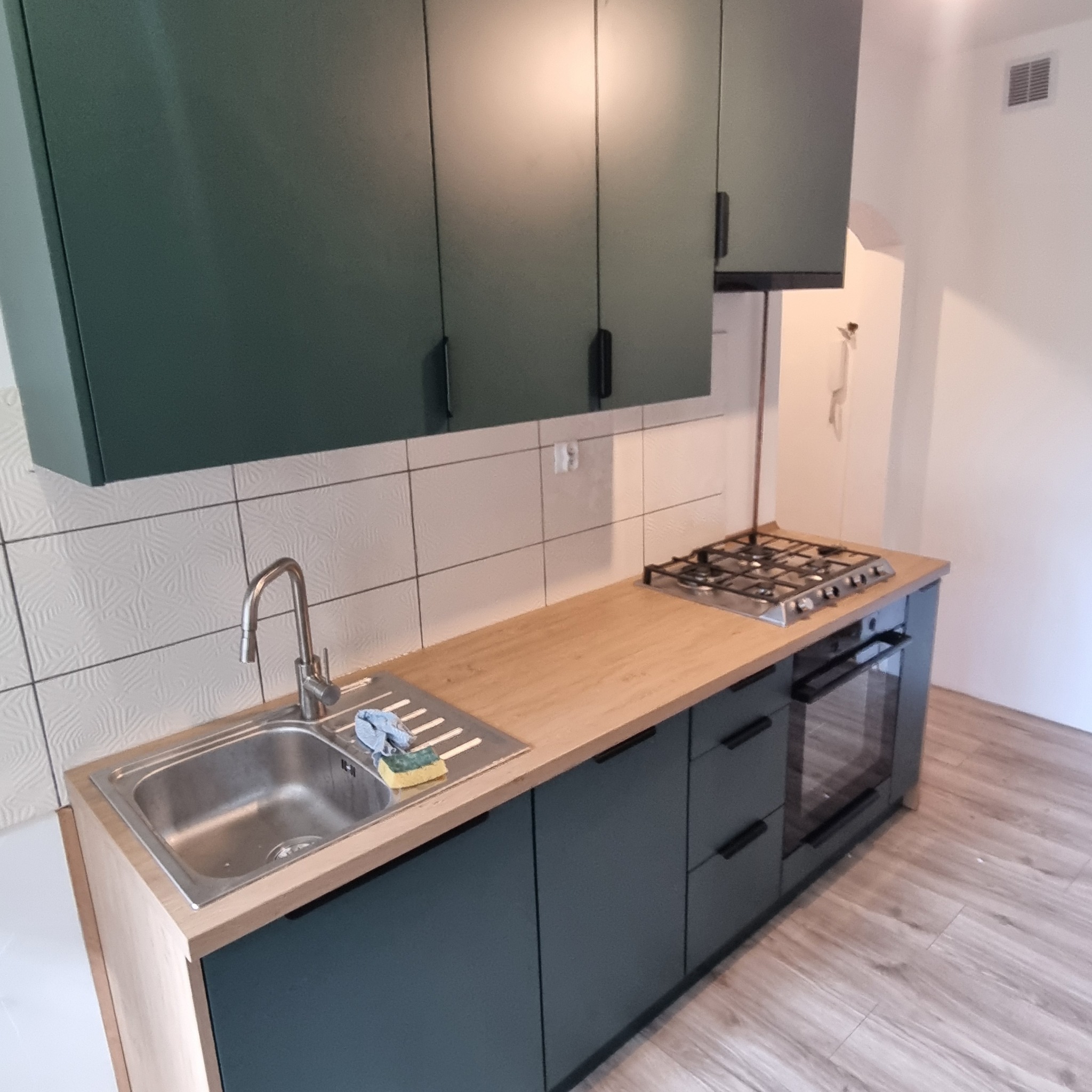 Nowoczesna kuchnia z ciemnozielonymi frontami, drewnianym blatem i białymi płytkami. Widoczny zlewozmywak, płyta gazowa i piekarnik. Ujęcie z perspektywy podłogi.