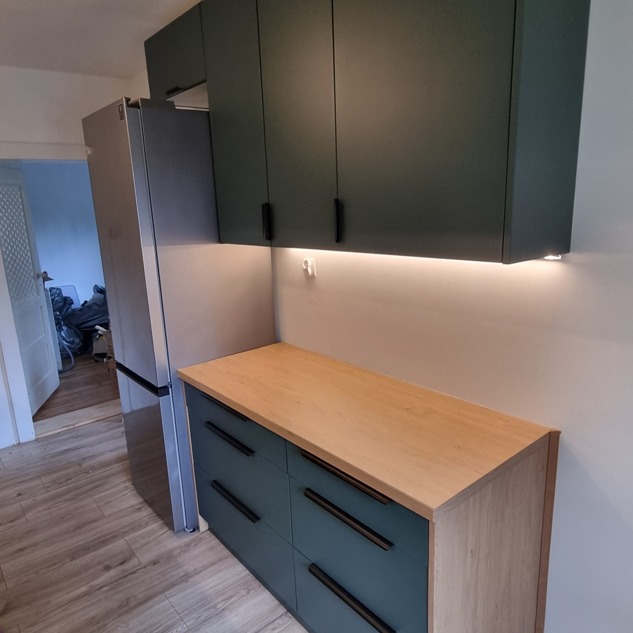 Nowoczesna kuchnia: ciemnozielone szafki wiszące i dolne z drewnianym blatem, oświetlenie LED podszafkowe, srebrna lodówka. Minimalistyczny design.