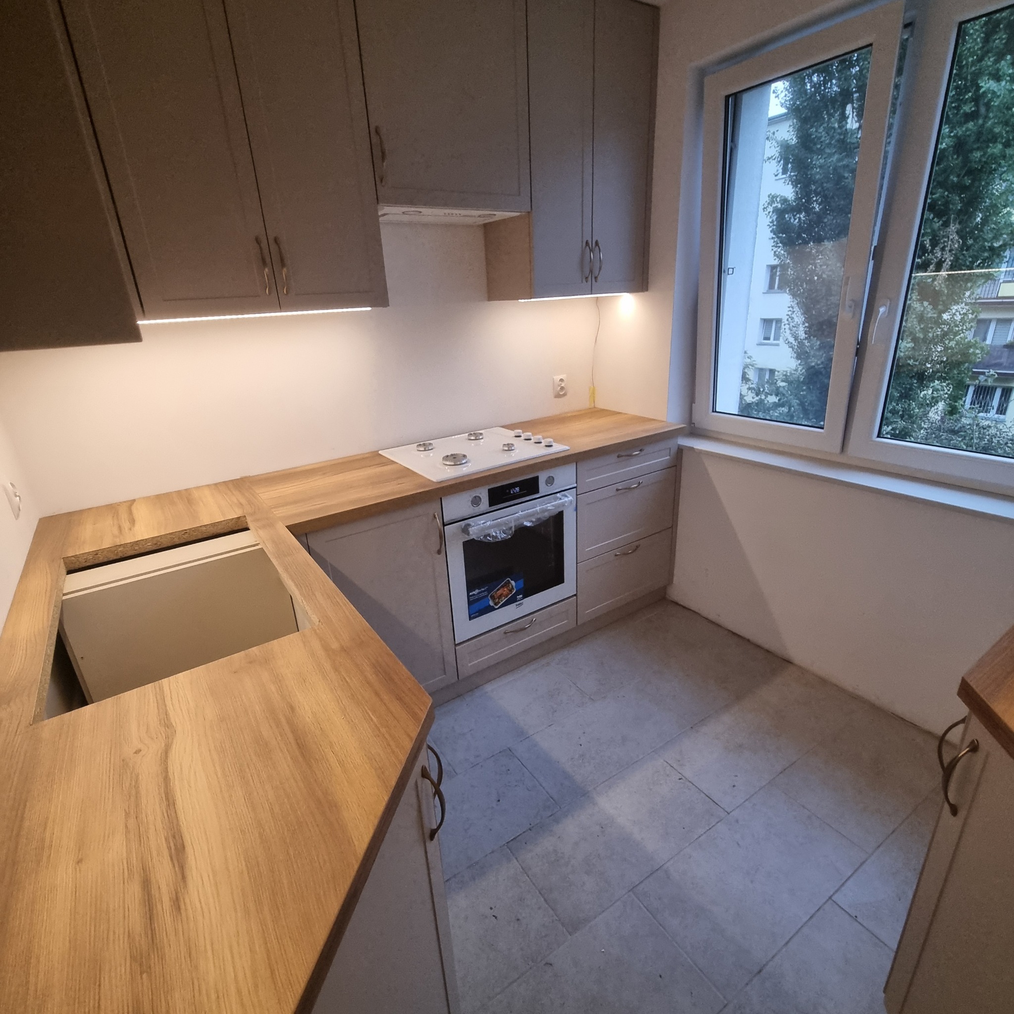 Jasna kuchnia z drewnianym blatem i szarymi szafkami, oświetlona listwą LED. Widok na okno z zielenią na zewnątrz, płytki na podłodze. Nowoczesny design.