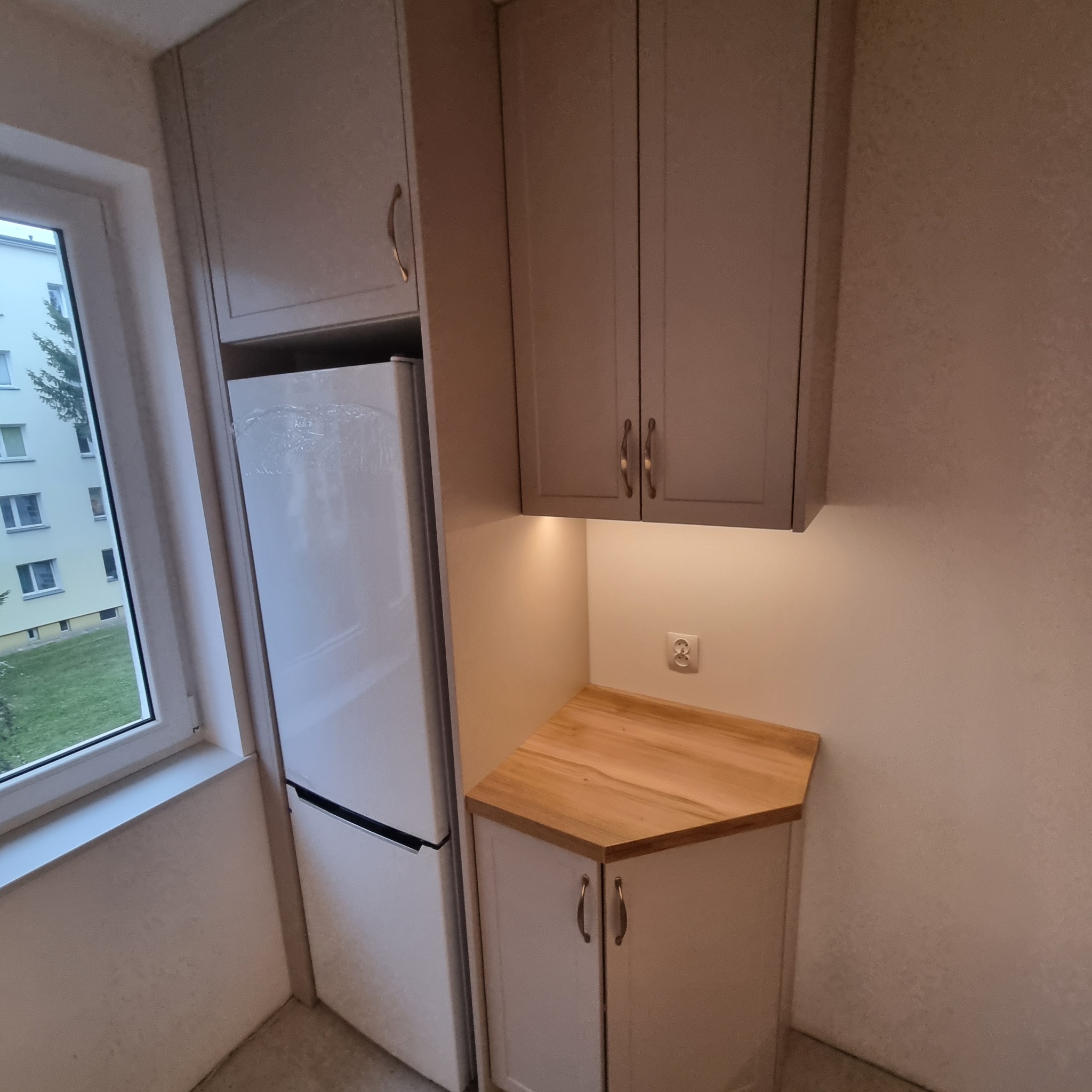 Jasna zabudowa kuchenna z drewnianym blatem i oknem z widokiem na zieleń. Uwagę zwracają metalowe uchwyty szafek i podświetlenie blatu.