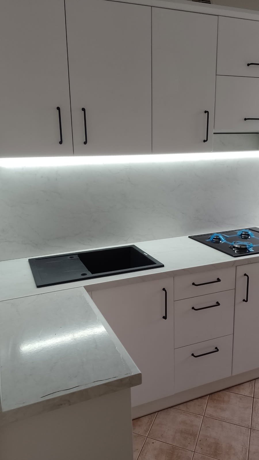 Biała kuchnia z czarnym zlewozmywakiem i płytą grzewczą, oświetlenie LED pod szafkami. Minimalistyczny design z marmurowym blatem i czarnymi uchwytami.