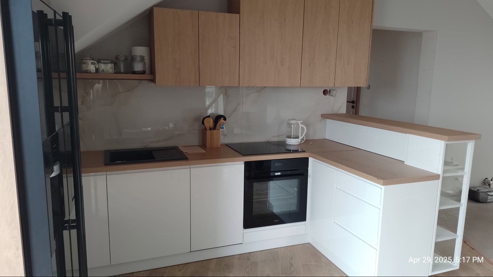 Nowoczesna kuchnia na wymiar z białymi frontami i drewnianymi blatami, wbudowany piekarnik i płyta indukcyjna. Uwagę zwraca skośny sufit i półka z dekoracjami.