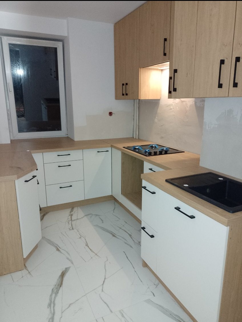 Jasna kuchnia z drewnianymi frontami i czarnymi uchwytami, marmurowe płytki podłogowe. Widoczne okno i fragment ściany w trakcie wykańczania.