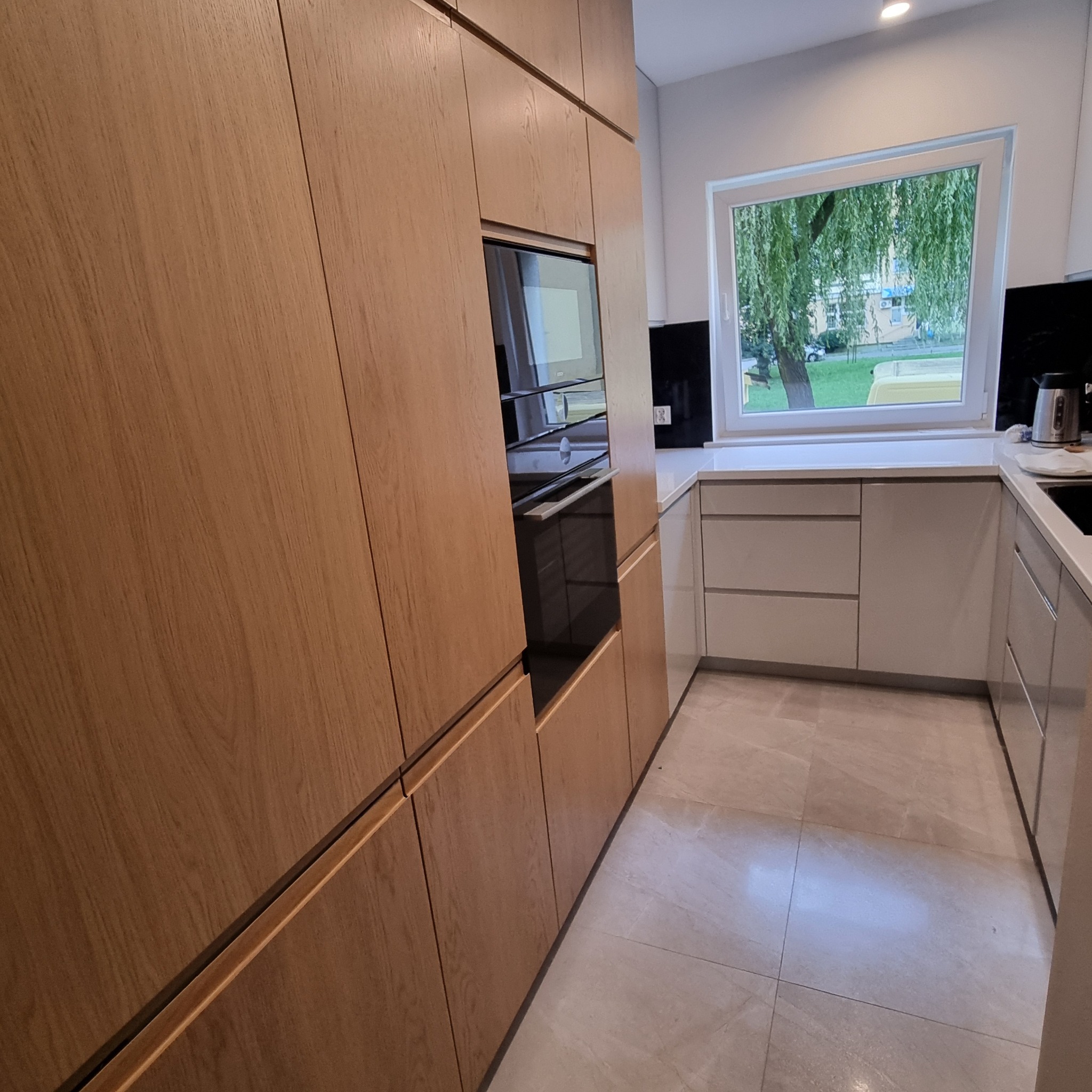 Nowoczesna kuchnia z drewnianymi frontami i wbudowanym piekarnikiem, widok na okno z zielenią, jasne płytki podłogowe.