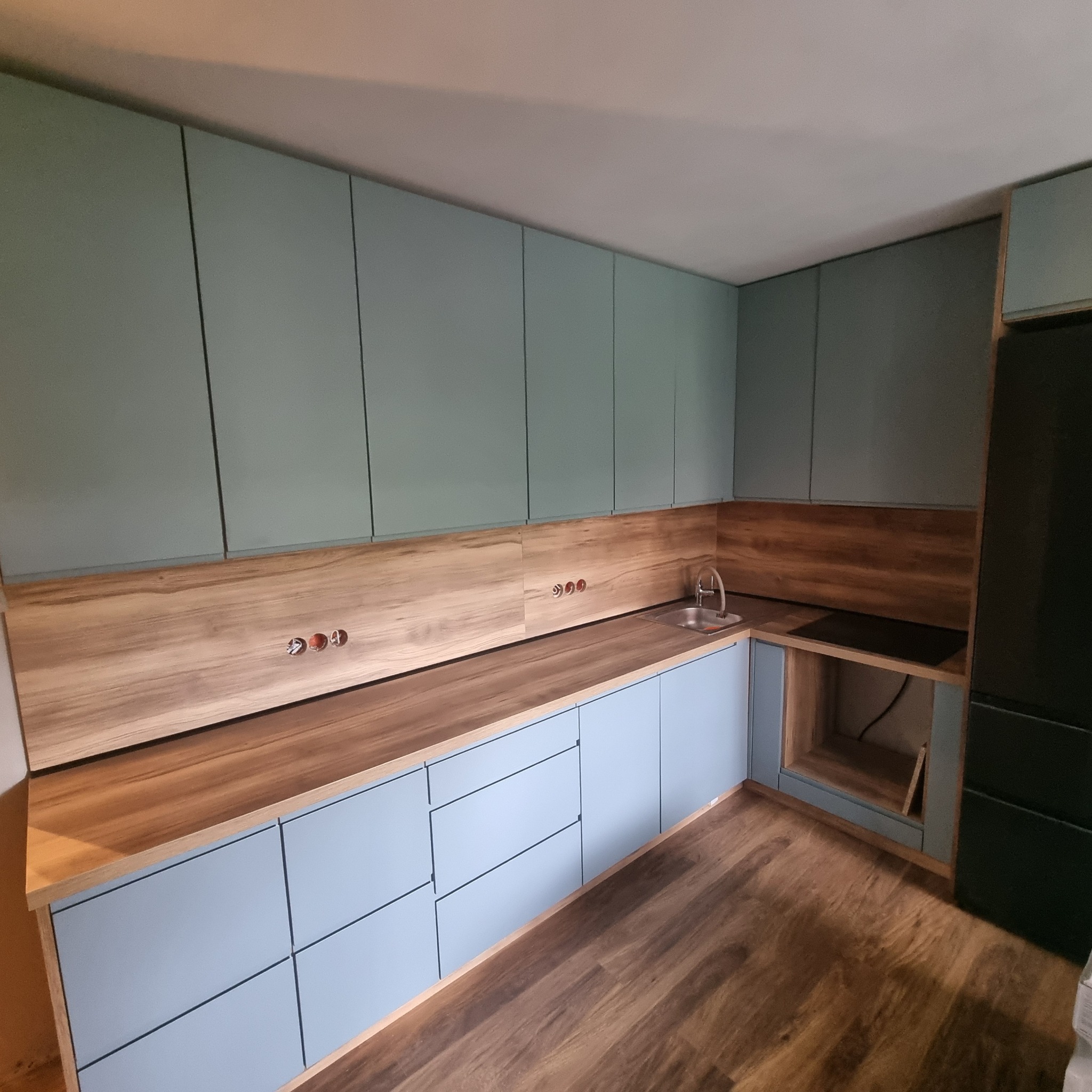 Jasna kuchnia z błękitnymi szafkami i drewnianym blatem. Widoczne gniazdka, zlew i płyta grzewcza. Podłoga drewniana, w tle czarna lodówka.