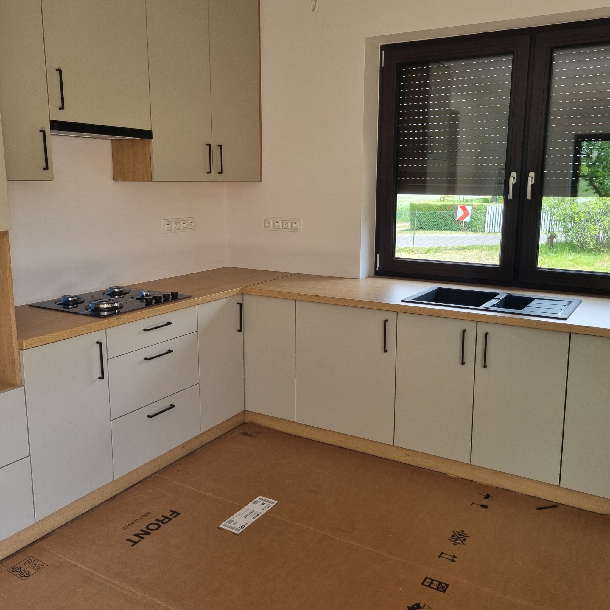 Jasna kuchnia z drewnianym blatem i czarnymi uchwytami, widok na okno z roletami. Na podłodze karton zabezpieczający. Minimalistyczny styl.