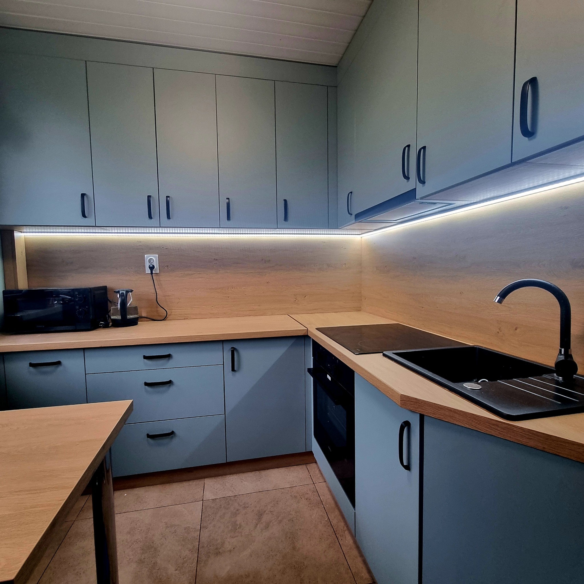 Nowoczesna kuchnia w odcieniach szarości z drewnianym blatem i oświetleniem LED. Widoczne szafki górne i dolne, zlew, płyta grzewcza i sprzęt AGD. Minimalistyczny design.
