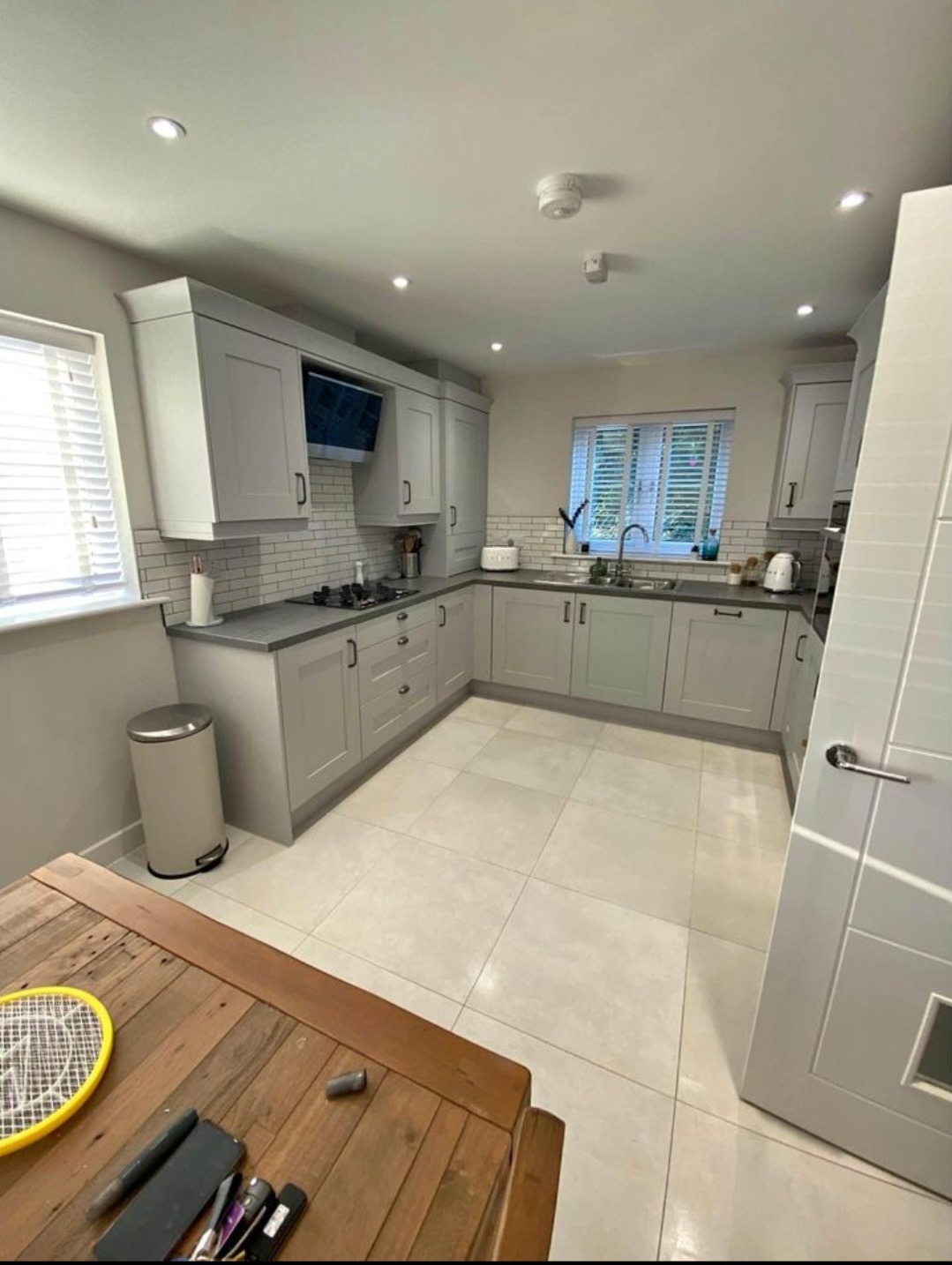 Jasna kuchnia w kształcie litery U z szarymi frontami, białymi płytkami na ścianie i drewnianym stołem z akcesoriami kuchennymi na pierwszym planie.