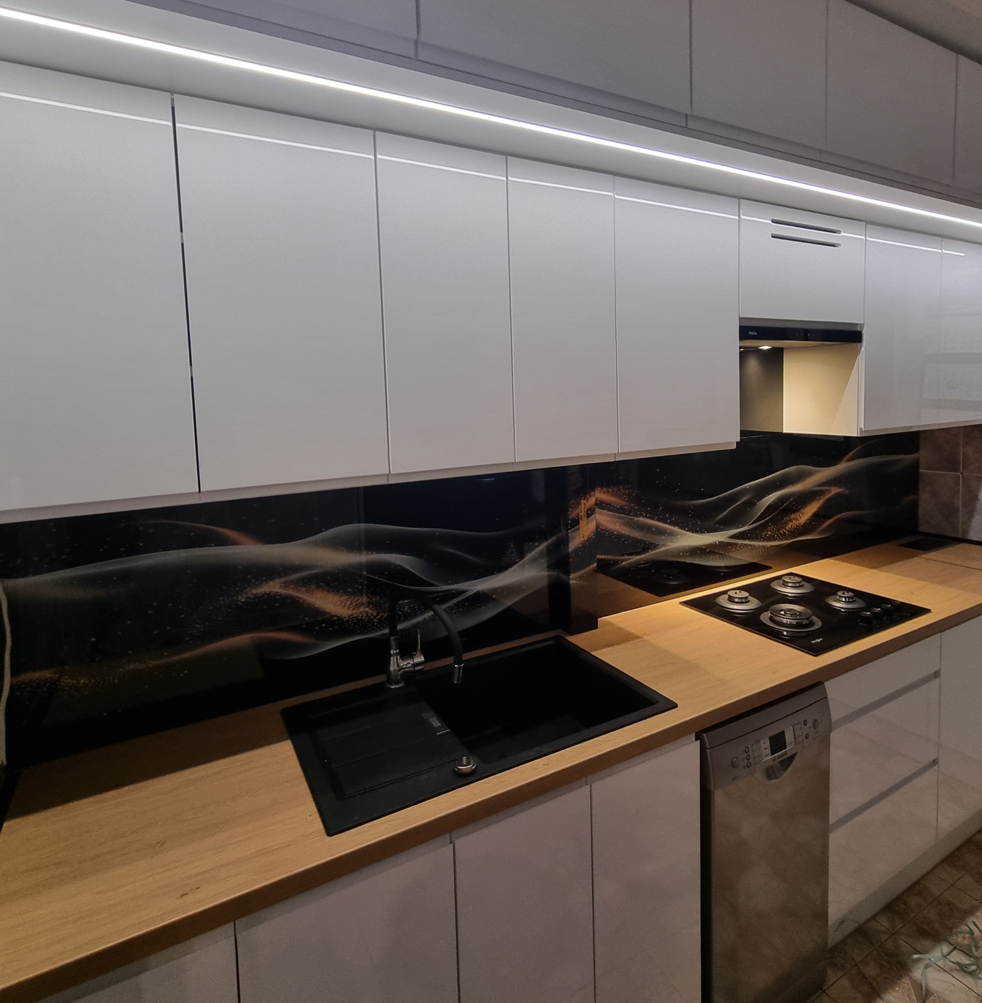 Nowoczesna kuchnia z białymi szafkami, drewnianym blatem i czarnym panelem ze złotym wzorem. Widoczny zlew, płyta grzewcza i oświetlenie LED pod szafkami.