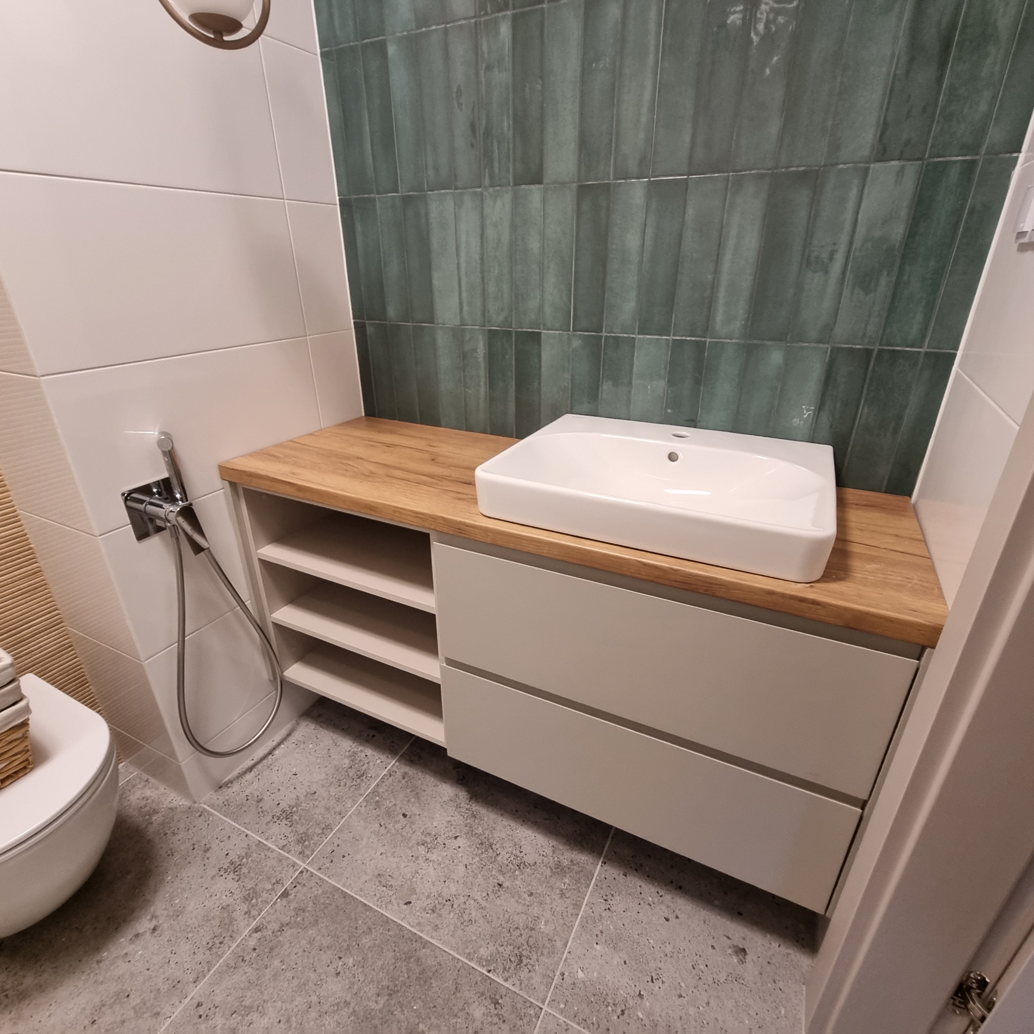 Nowoczesna szafka łazienkowa na wymiar z drewnianym blatem i białą umywalką. Na ścianie zielona, pionowa glazura. Widoczne elementy toalety i bidetka.