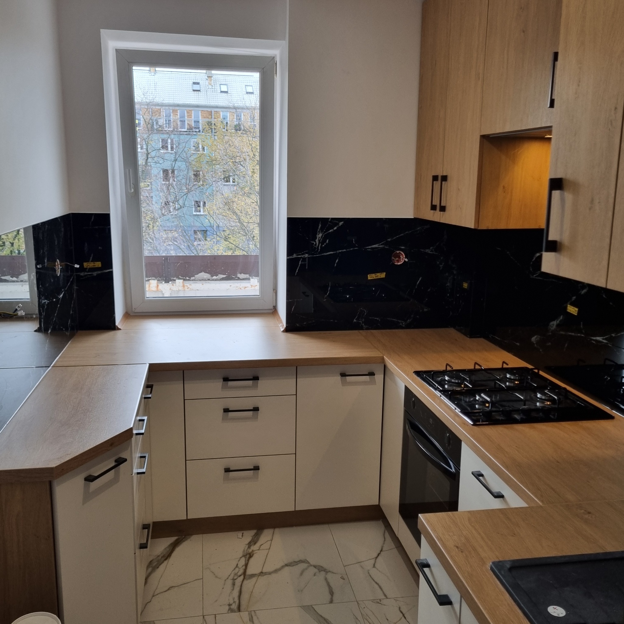 Jasna kuchnia z drewnianymi blatami i czarnym panelem nad blatem, widok na okno z zabudową miejską w tle. Minimalistyczne uchwyty szafek.