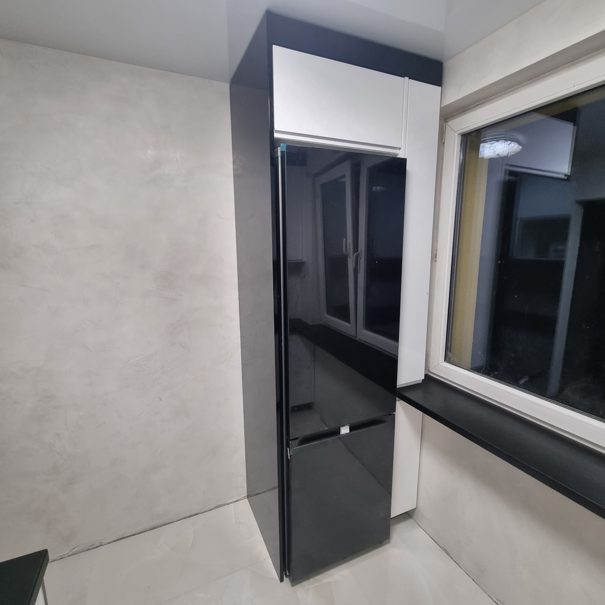 Czarna lodówka w zabudowie kuchennej z białymi szafkami wiszącymi, widok z boku na tle szarej ściany i okna. Nowoczesny design.