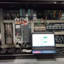 Otwarta szafa sterownicza z widocznymi modułami, okablowaniem i laptopem Dell z uruchomionym oprogramowaniem diagnostycznym, podłączonym do szafy kablem USB.