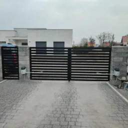 Nowoczesna, pozioma brama wjazdowa z automatyką, zamontowana na kamiennych słupkach, z widocznym podjazdem z kostki brukowej i fragmentem domu w tle.
