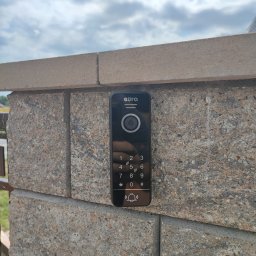 OMIautomatyka - Czarny panel wideodomofonu Eura z klawiaturą numeryczną zamontowany na szarym, kamiennym słupku ogrodzeniowym na tle pochmurnego nieba.