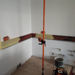 OMIautomatyka - Montaż dekoracyjnej ściany z cegły w pomieszczeniu z użyciem poziomicy laserowej, widoczne puszki elektryczne i przygotowane instalacje wodne.