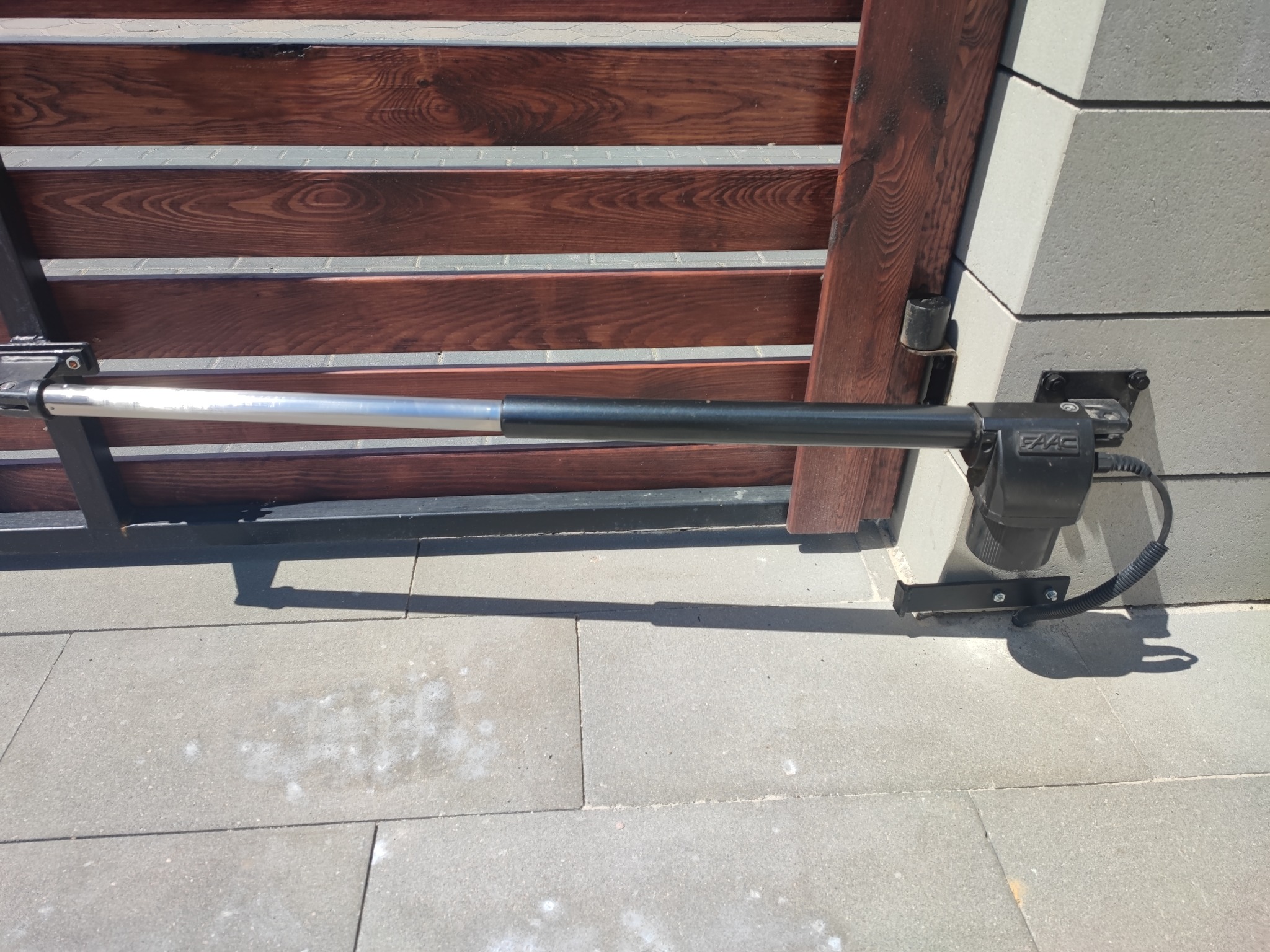 Siłownik bramy automatycznej FAAC zamontowany na murku z szarej kostki brukowej, widoczny fragment drewnianej bramy z poziomych desek.