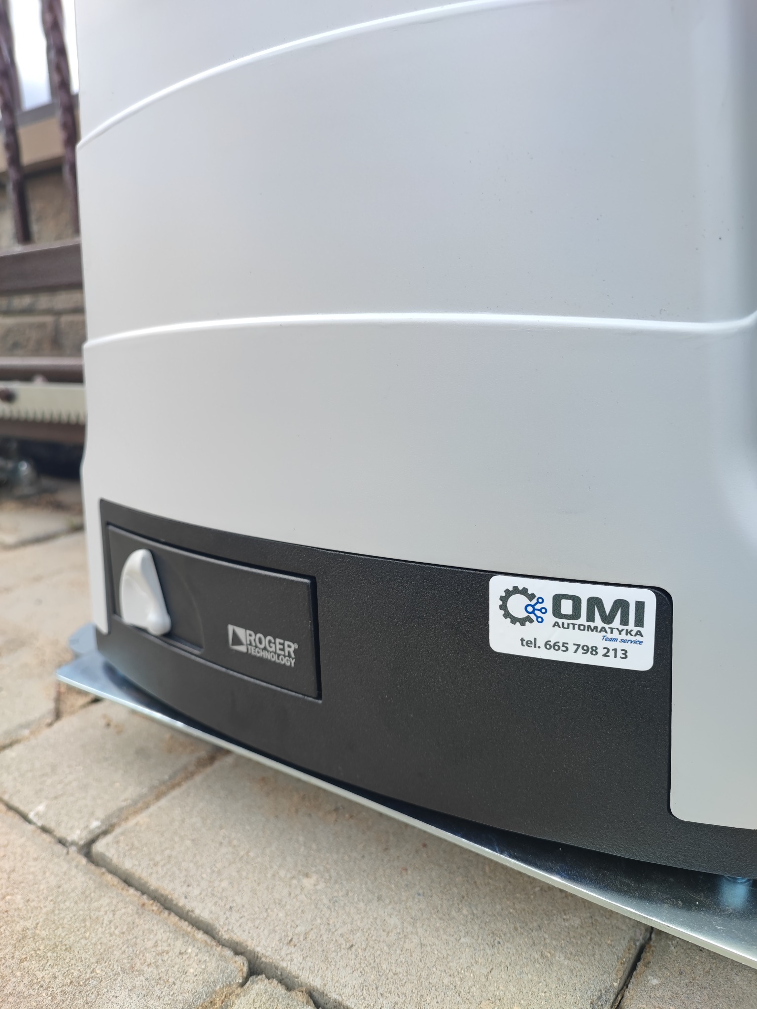 Szary napęd do bramy firmy Roger Technology, zamontowany na metalowej podstawie, z naklejką firmy OMI Automatyka Team Service z numerem telefonu.
