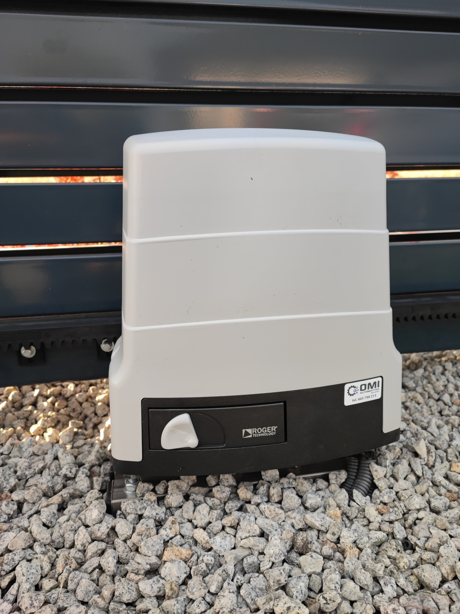 Szary automat bramy przesuwnej Roger Technology zamontowany na szarej bramie i podłożu z drobnych kamieni.