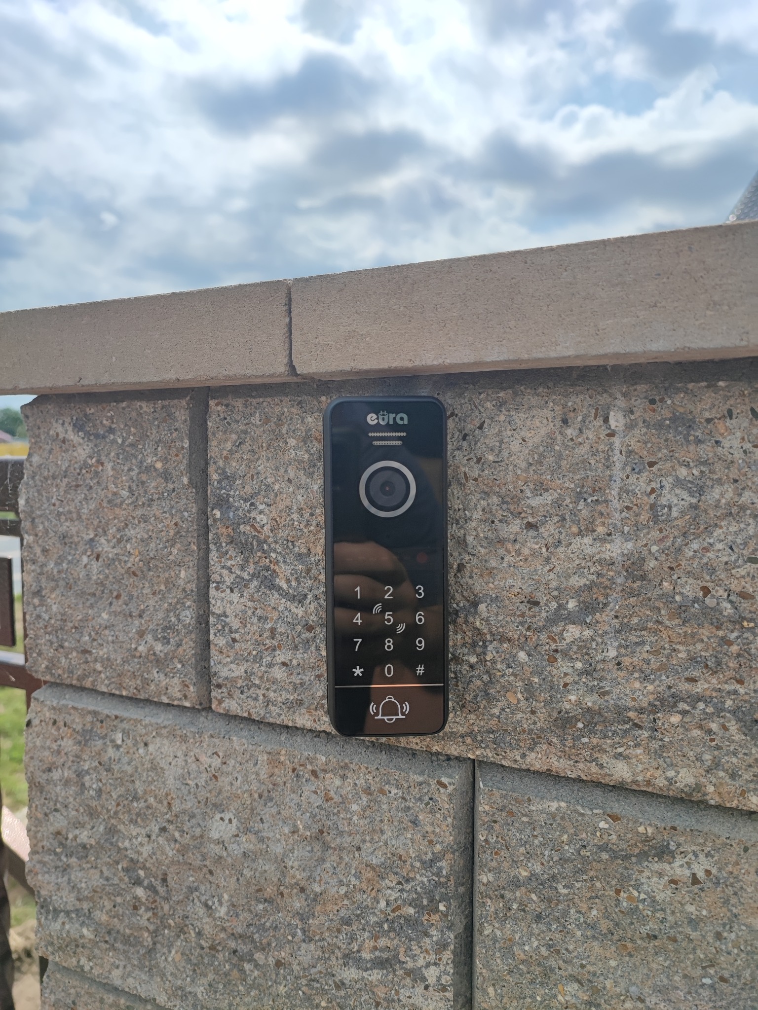 Czarny panel wideodomofonu Eura z klawiaturą numeryczną zamontowany na szarym, kamiennym słupku ogrodzeniowym na tle pochmurnego nieba.