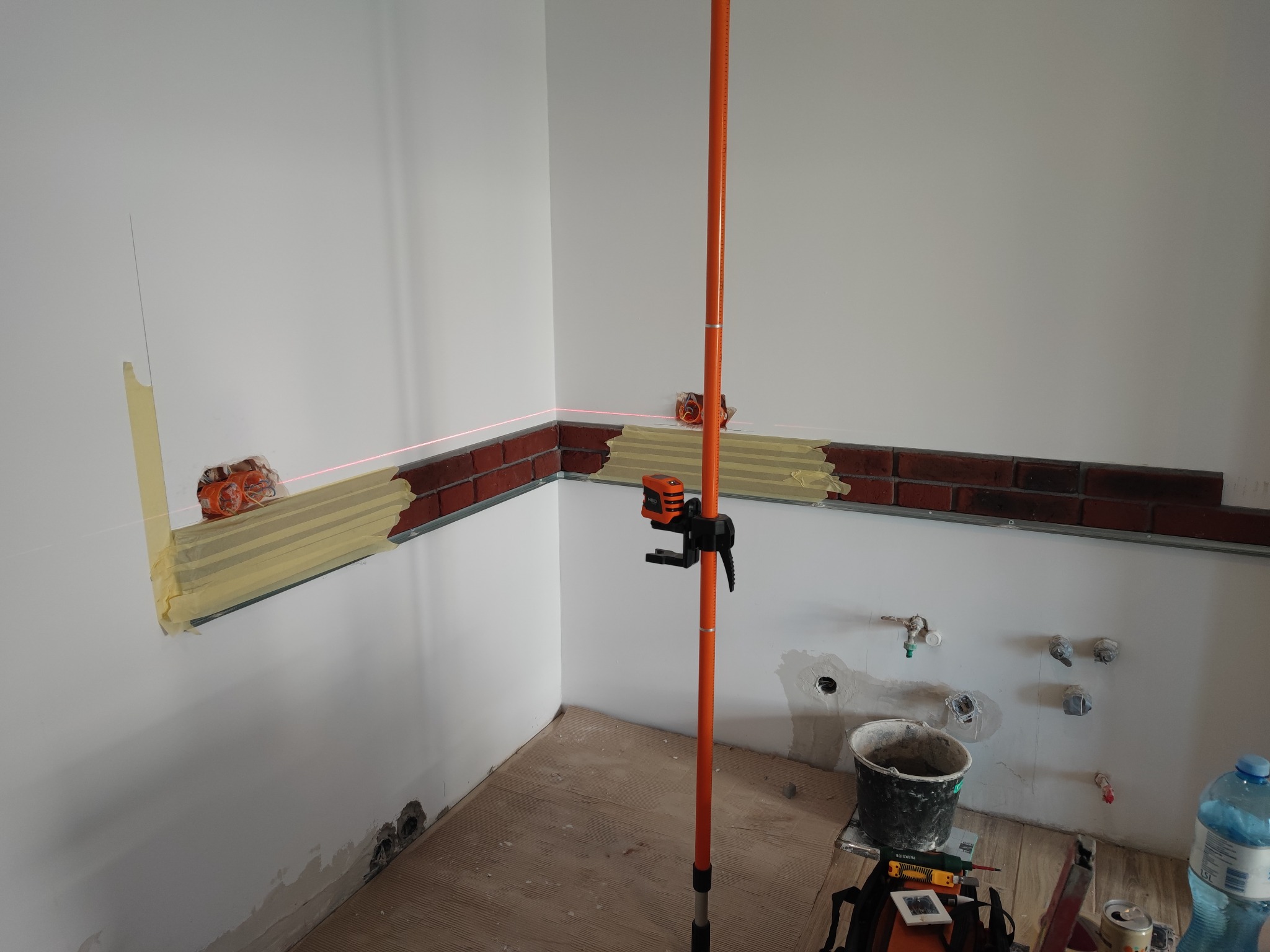 Montaż dekoracyjnej ściany z cegły w pomieszczeniu z użyciem poziomicy laserowej, widoczne puszki elektryczne i przygotowane instalacje wodne.
