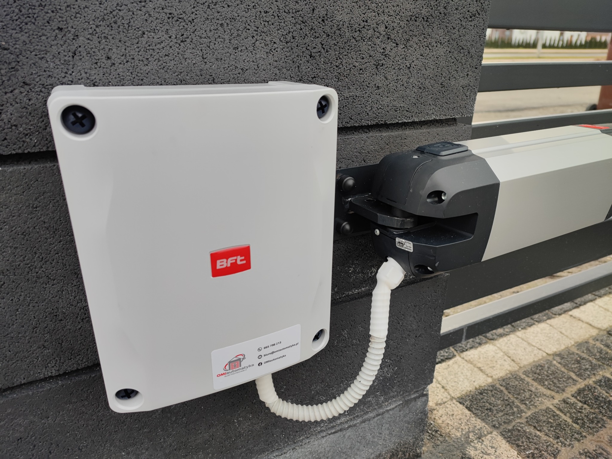 Zamontowany napęd bramy BFT z białą obudową sterującą i elastycznym przewodem ochronnym, przymocowany do szarego muru z fakturą, widok z bliska.