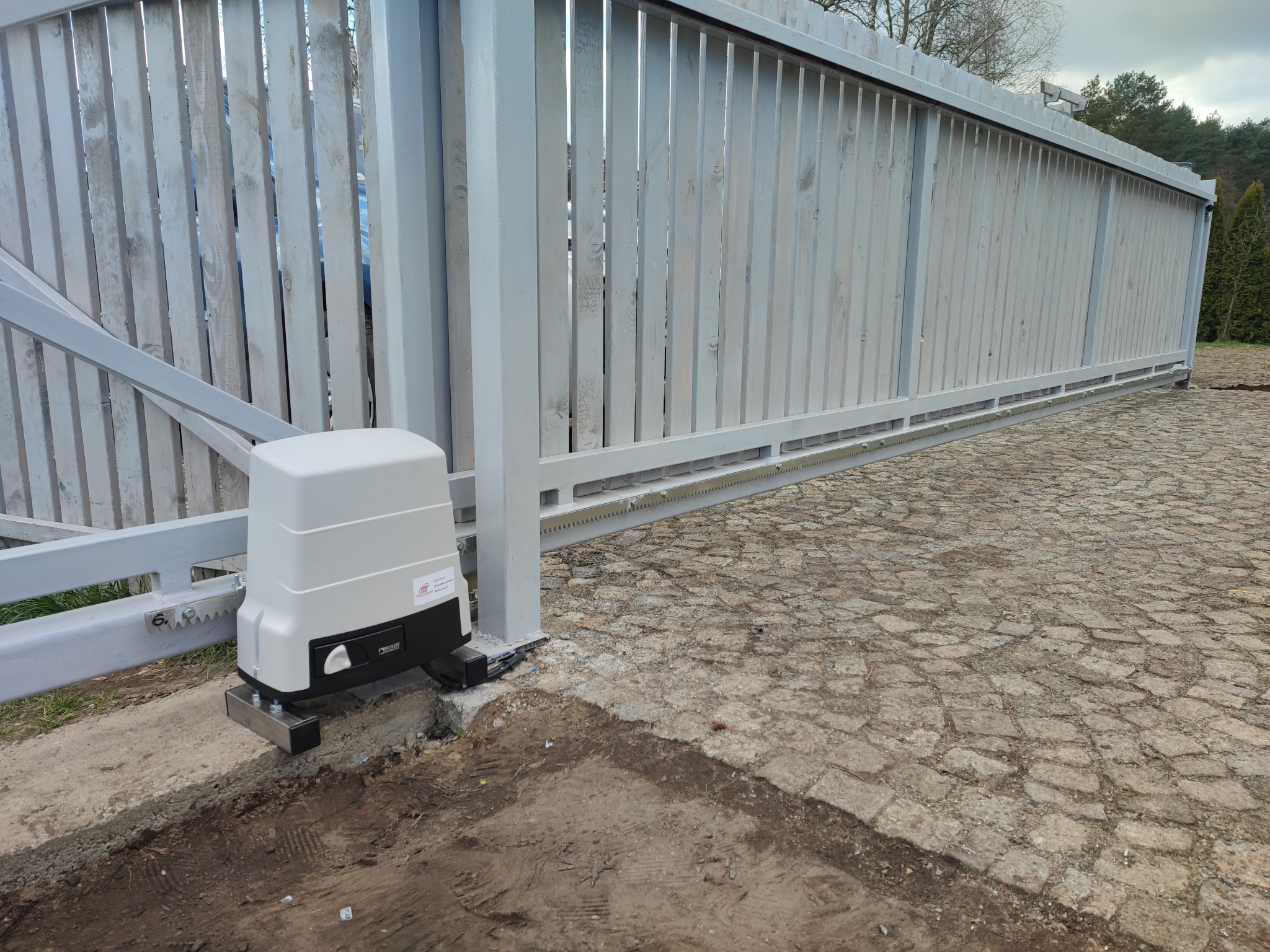 Zamontowany napęd do przesuwnej bramy posesyjnej z widoczną zębatką, szarą drewnianą bramą oraz brukowanym podjazdem.