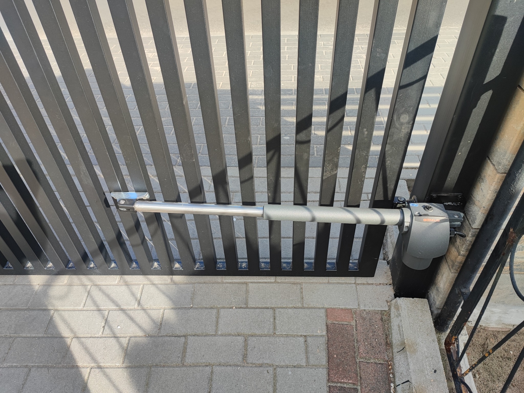 Szczegółowe ujęcie siłownika automatyki bramy skrzydłowej, widoczna szara obudowa mechanizmu i wysuwany teleskopowy element mocowany do czarnego, pionowo żebrowanego skrzydła bramy, na tle kostki...