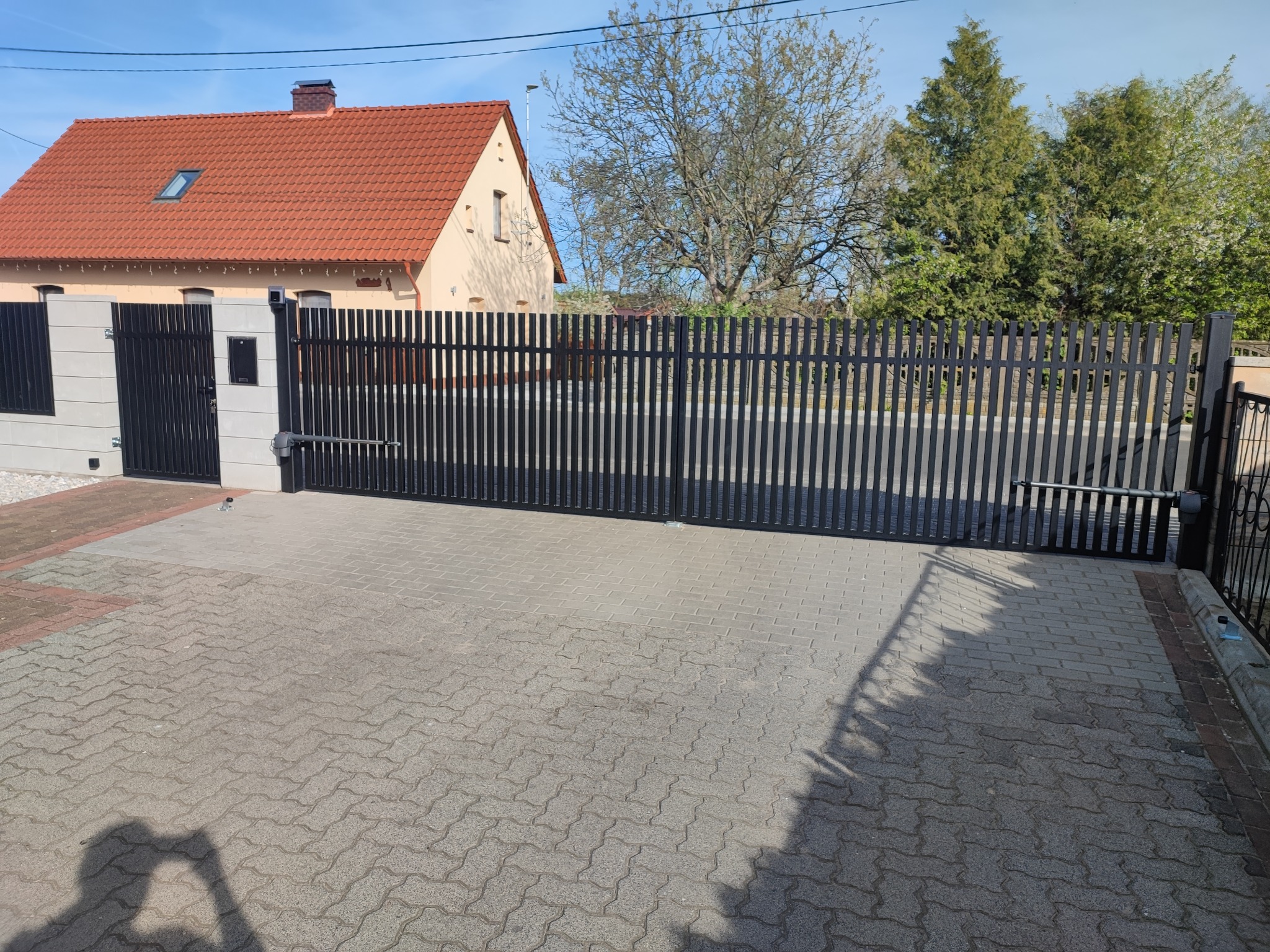 Czarna, nowoczesna brama przesuwna z automatyką, widoczna na tle domu z czerwoną dachówką i brukowanym podjazdem w słoneczny dzień.