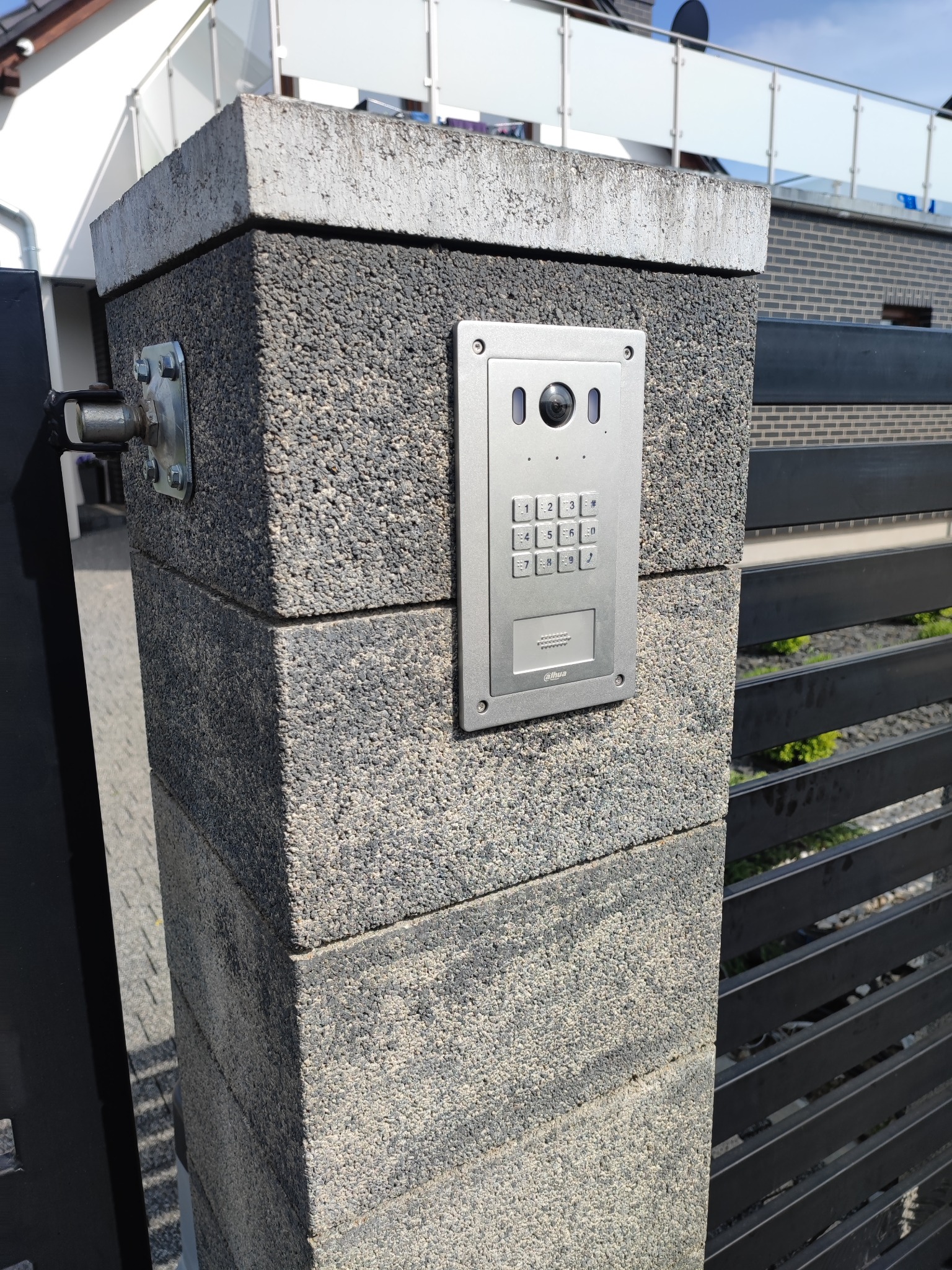 Szary domofon z klawiaturą numeryczną i kamerą zamontowany na słupku ogrodzeniowym z szarego betonu architektonicznego, widoczne czarne przęsła ogrodzenia.