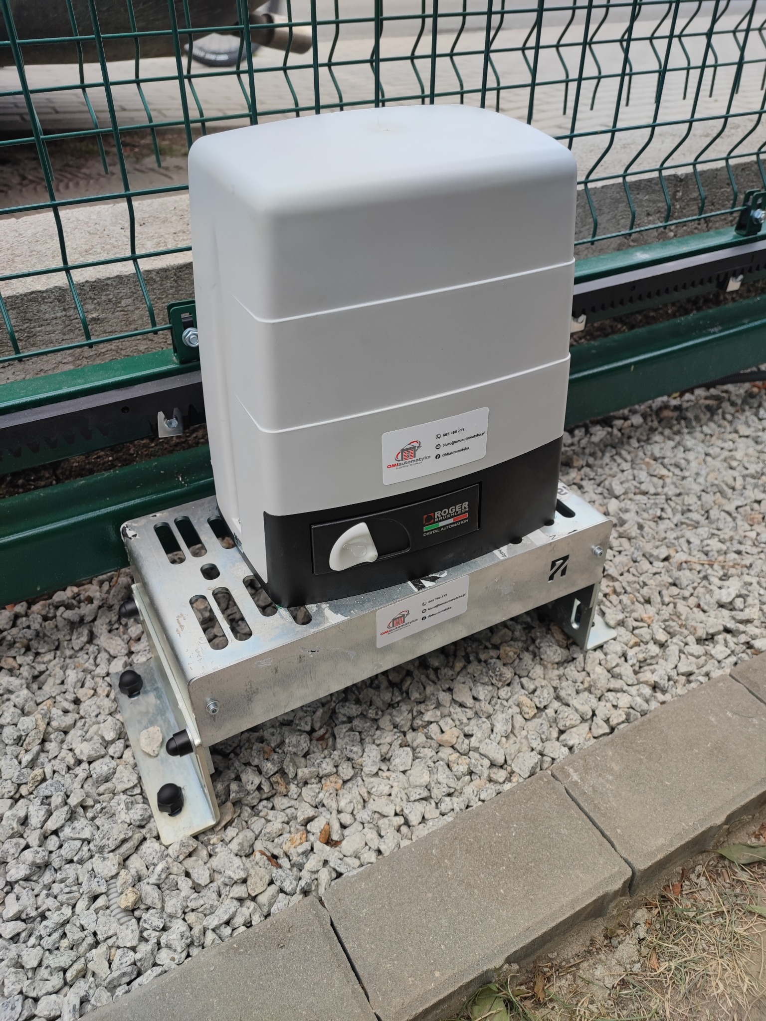Zamontowany napęd do bramy przesuwnej marki Roger na metalowej podstawie, widoczny mechanizm zębaty i fragment ogrodzenia panelowego.