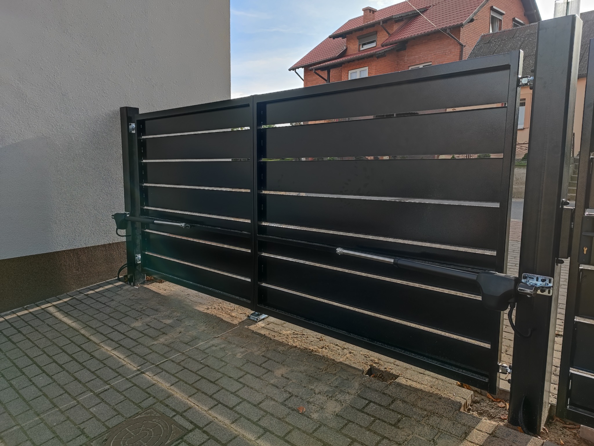 Nowoczesna, czarna brama przesuwna z poziomymi, chromowanymi akcentami i siłownikami automatyki, zamontowana na kostce brukowej przed budynkiem mieszkalnym.